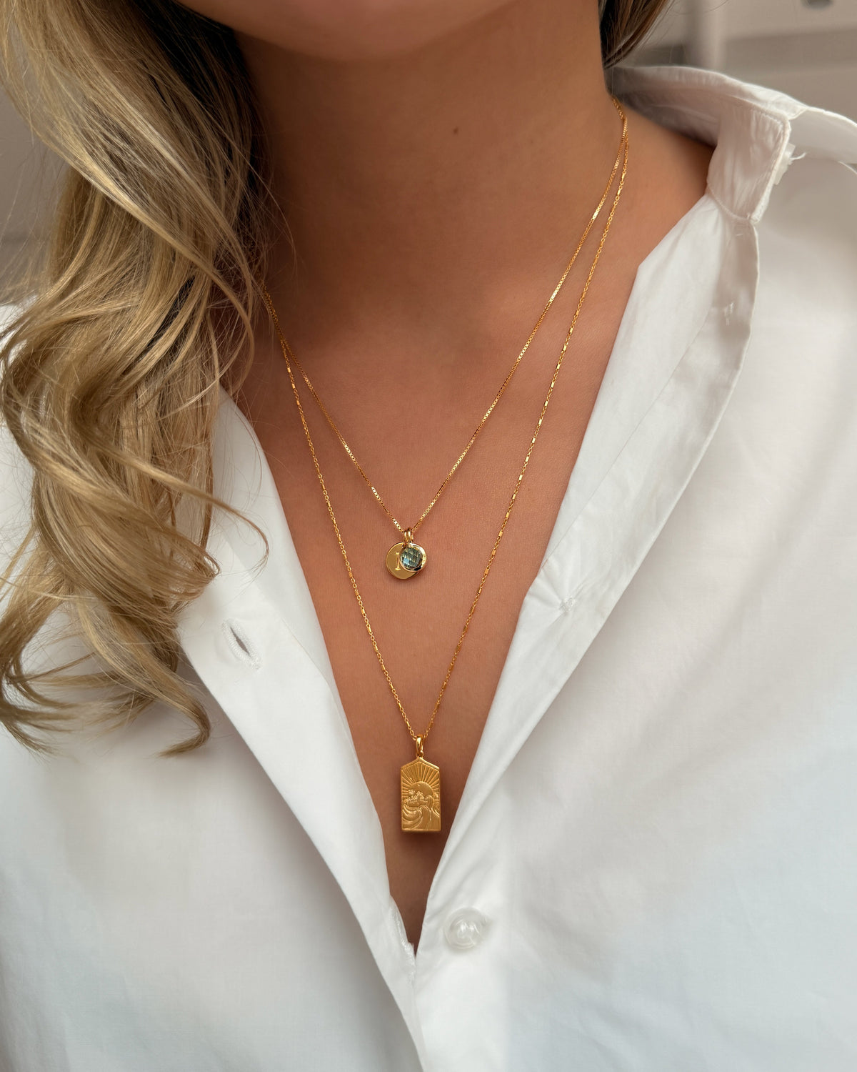 Two gold necklaces: the shorter features a September birthstone pendant set with blue topaz, while the longer is adorned with a rectangular pendant featuring a sun design.