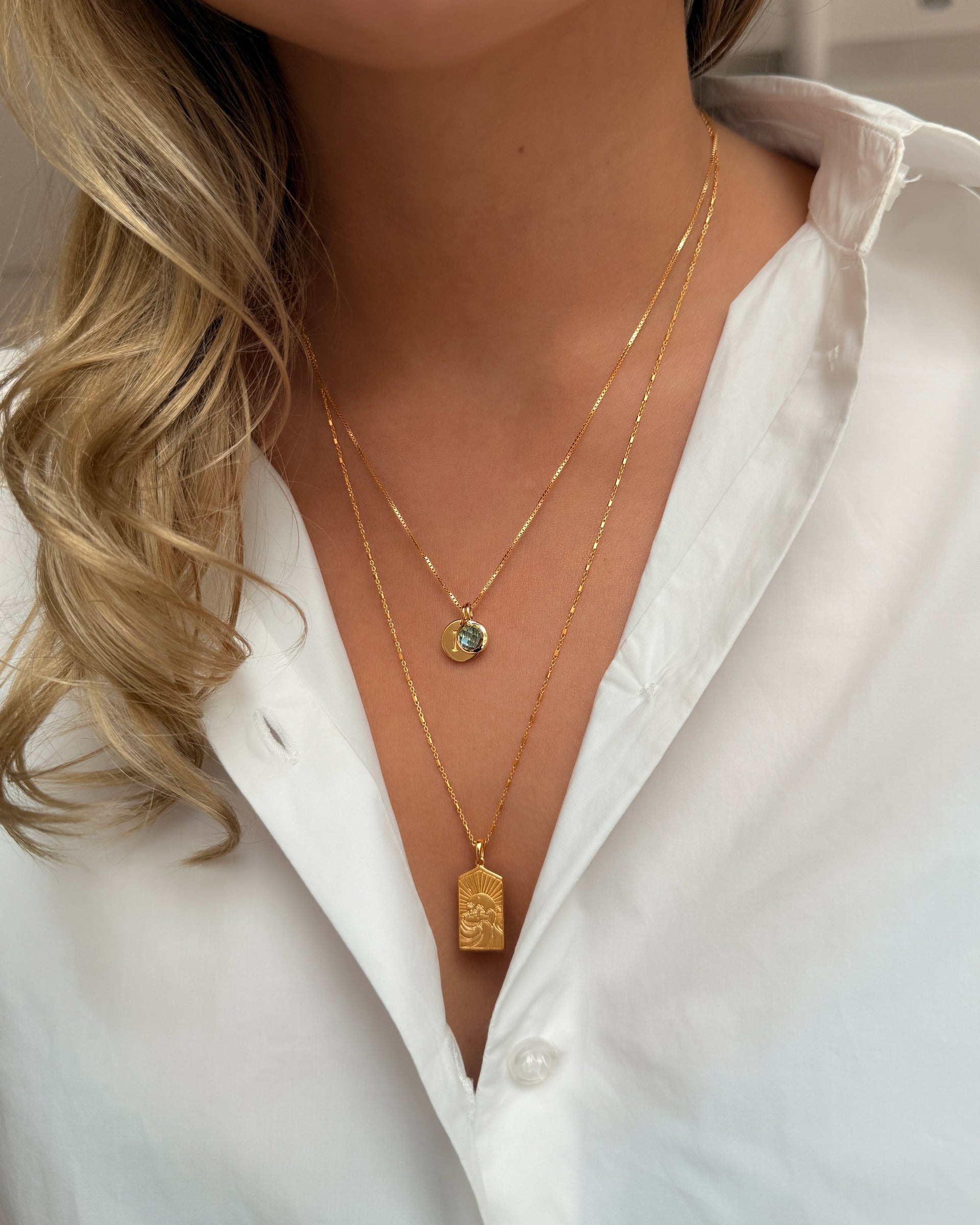 Two gold necklaces: the shorter features a September birthstone pendant set with blue topaz, while the longer is adorned with a rectangular pendant featuring a sun design.