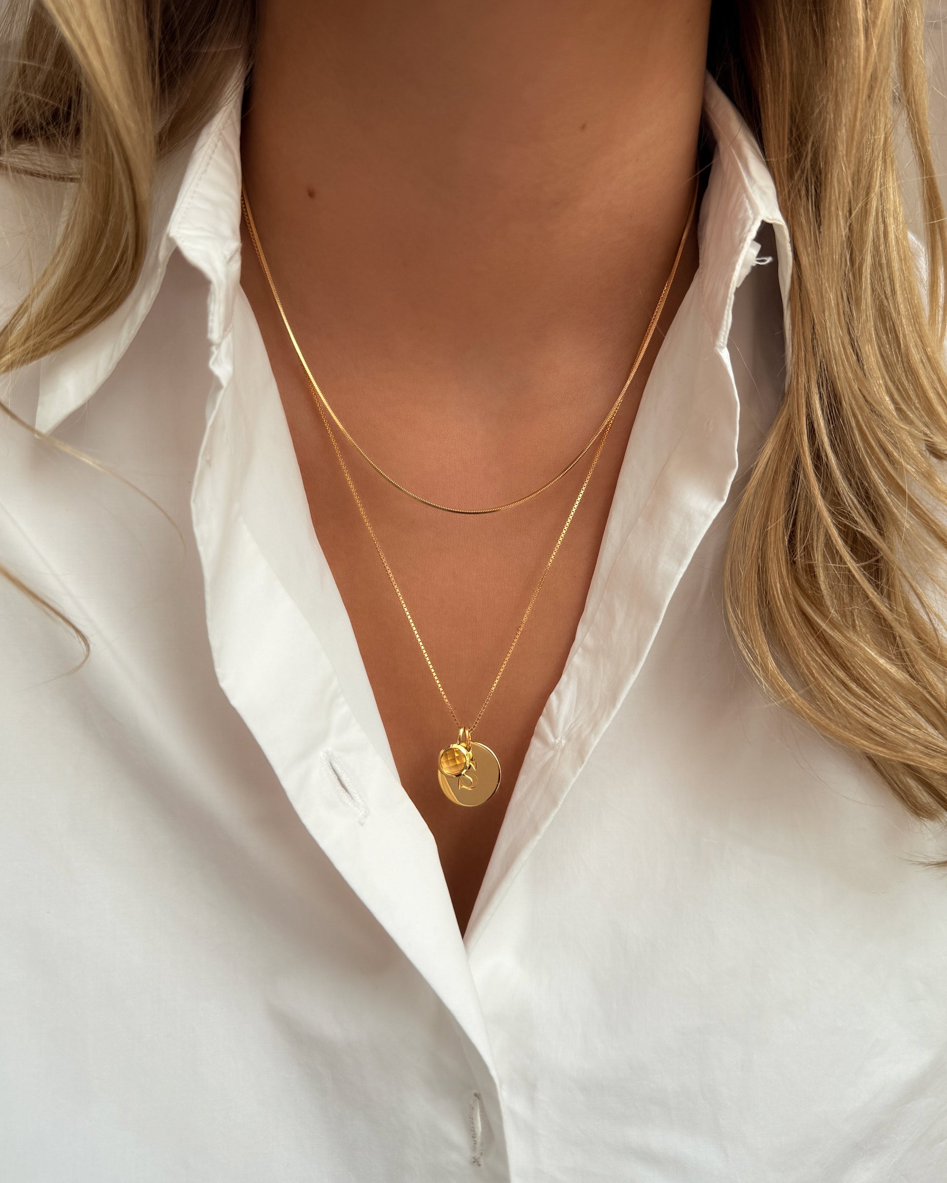 Two layered gold necklaces: a longer chain featuring a November birthstone citrine pendant and a round letter charm, paired with a shorter gold snake chain.