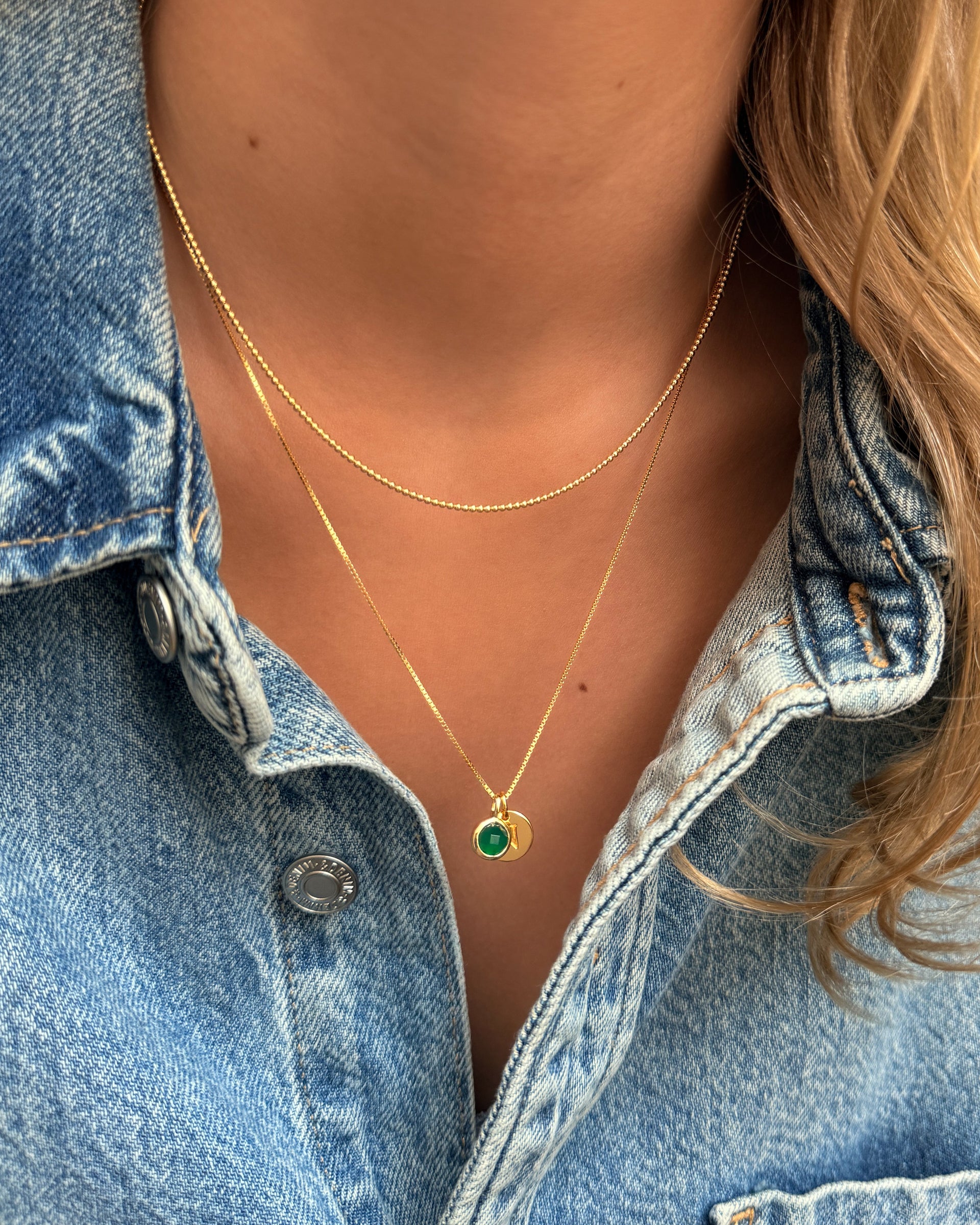 A gold pendant necklace featuring the May birthstone green onyx, layered with a gold chain and a letter charm, paired with a second gold beaded necklace.