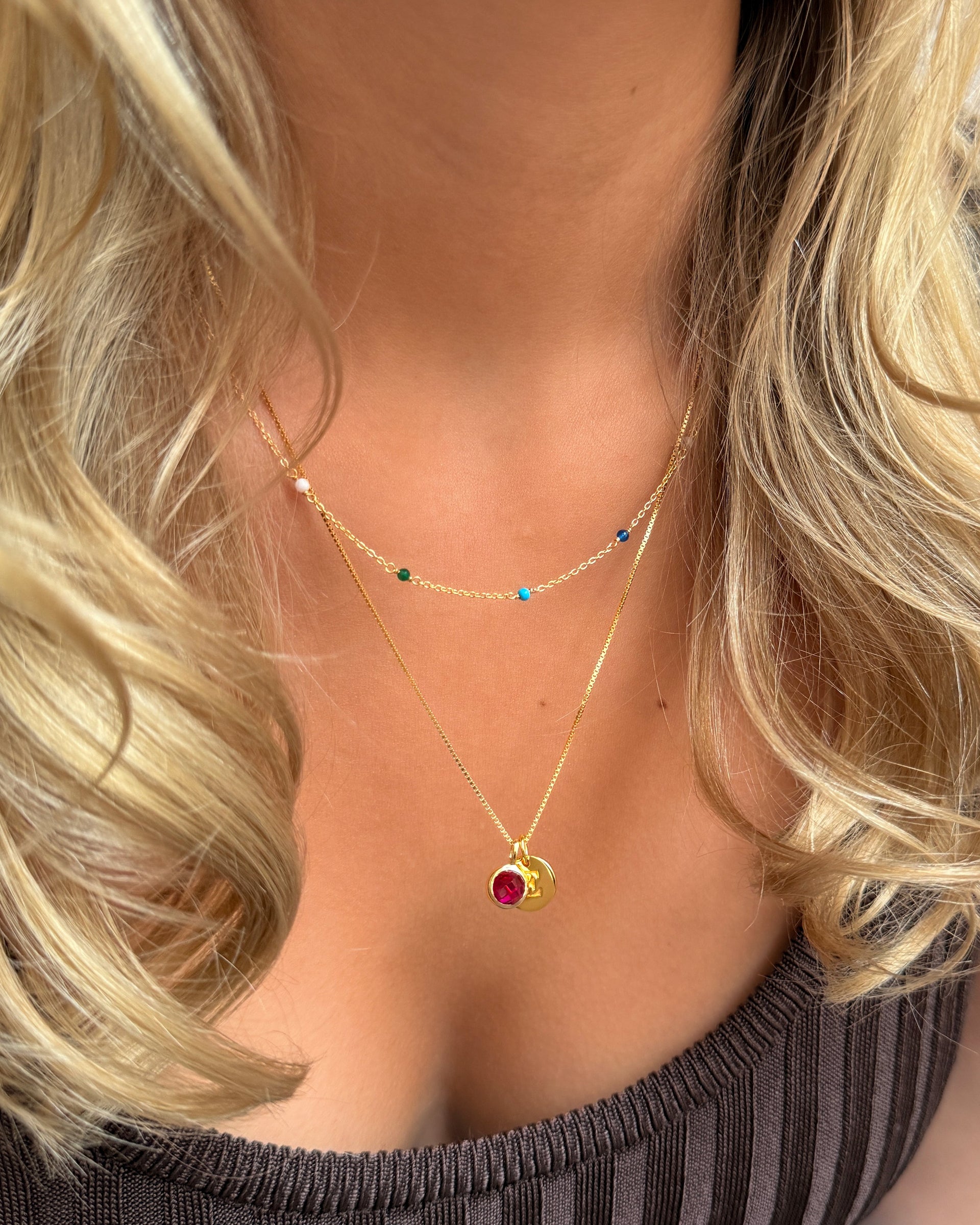 The gold Beloved Birthstone July pendant with a red ruby, paired with a small gold letter pendant on a gold chain, and layered with a second, shorter gold chain featuring small colorful beads.
