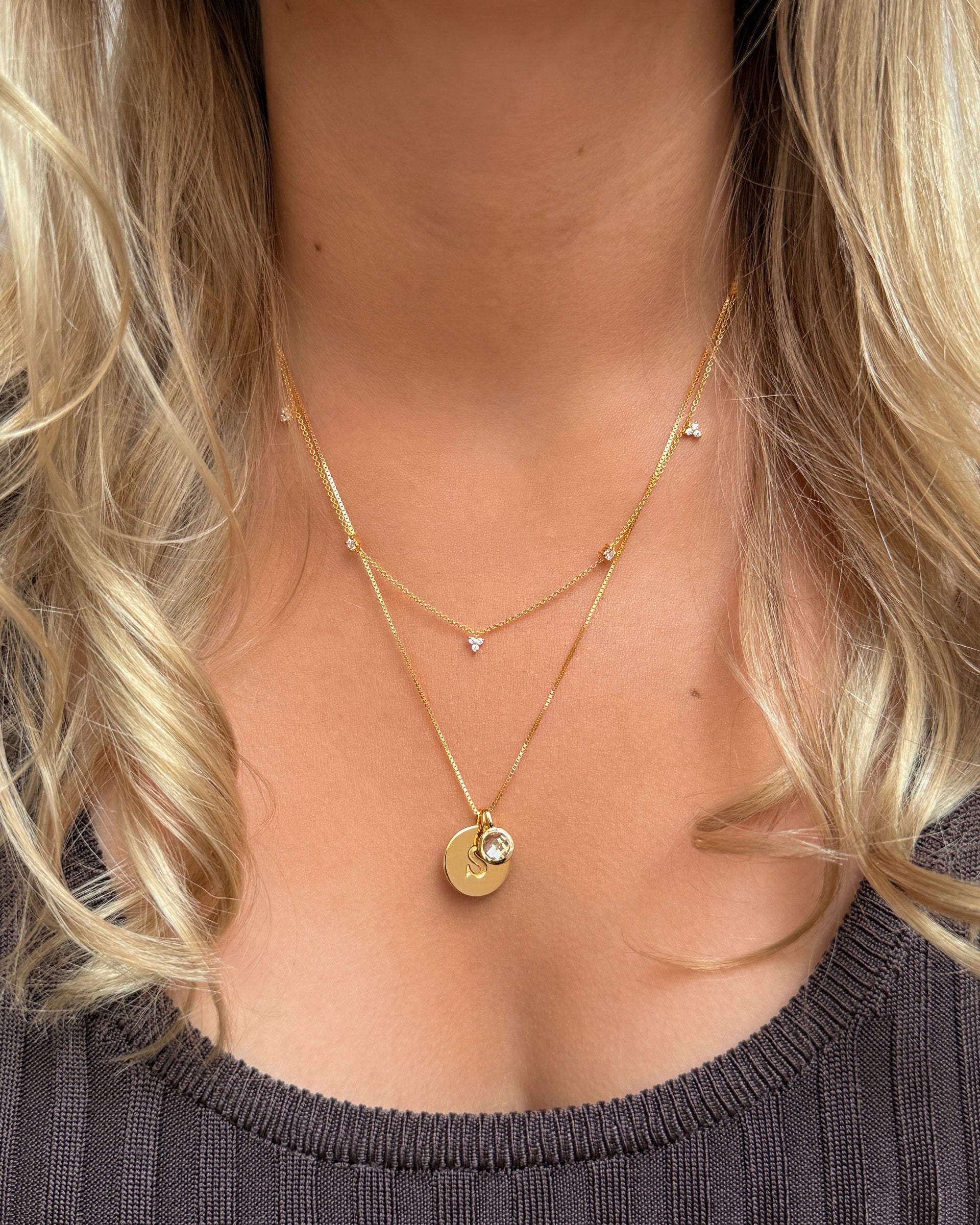A layered necklace set featuring an April birthstone pendant in gold with a white topaz, paired with a gold initial S pendant on a box chain. The set is completed with a shorter, delicate gold necklace adorned with small crystals.