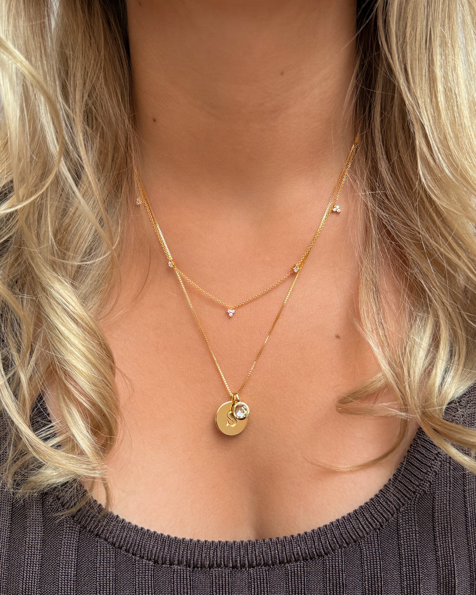 A layered necklace set featuring an April birthstone pendant in gold with a white topaz, paired with a gold initial S pendant on a box chain. The set is completed with a shorter, delicate gold necklace adorned with small crystals.