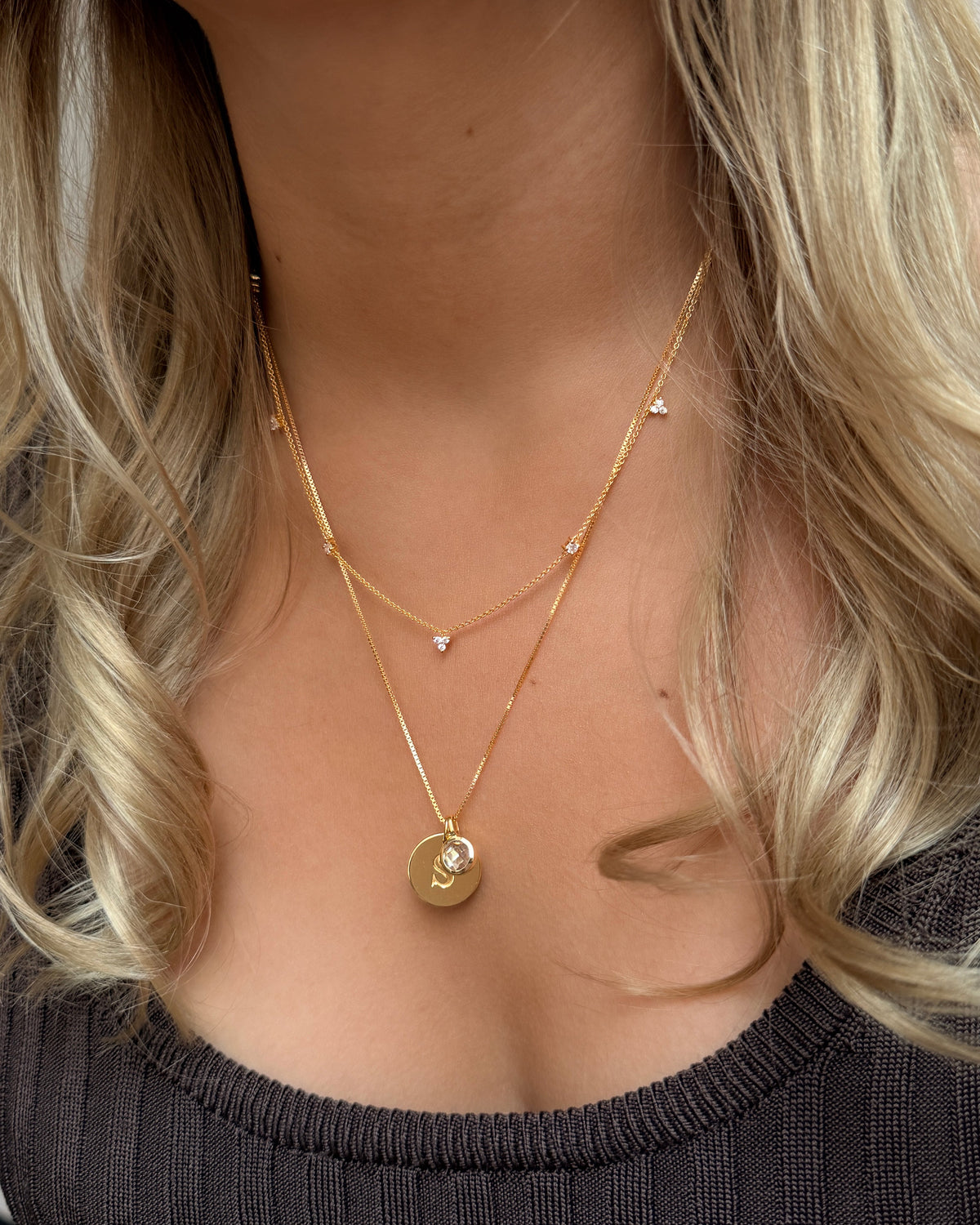 Two layered gold necklaces: a longer chain featuring an April birthstone white topaz pendant layered over a round initial charm, paired with a shorter delicate gold chain with small stones.