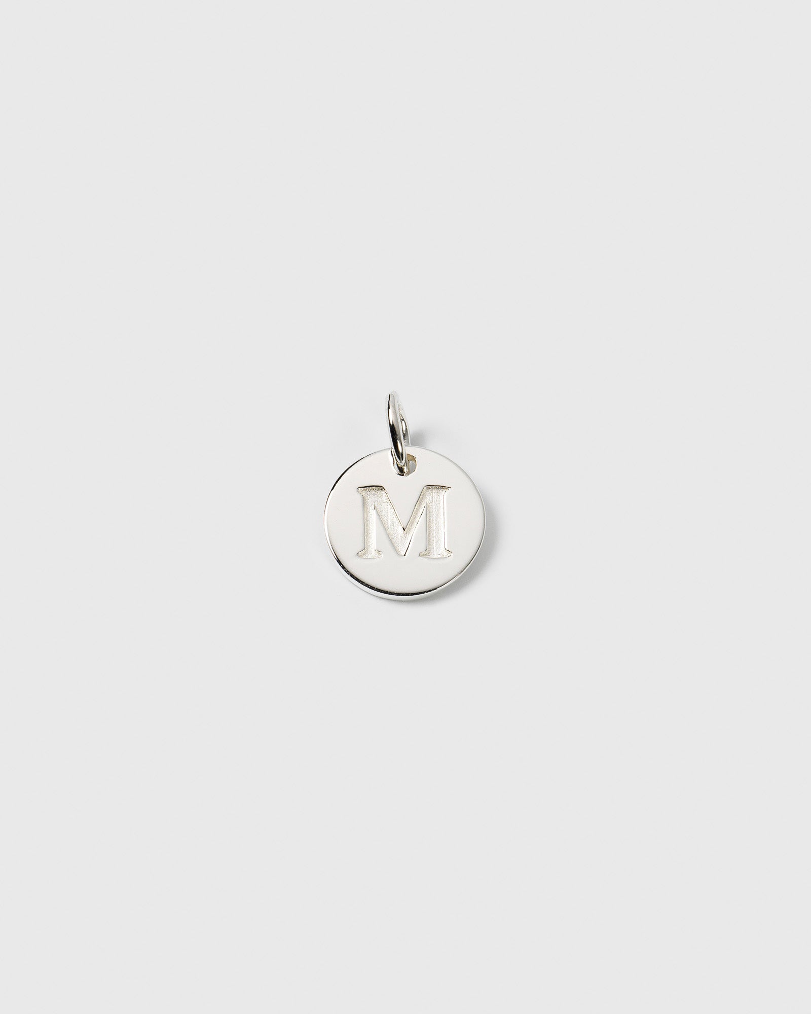 A product shot of the Beloved Mini Letter M charm in silver, a small, circular pendant with a capital M engraved on its surface.
