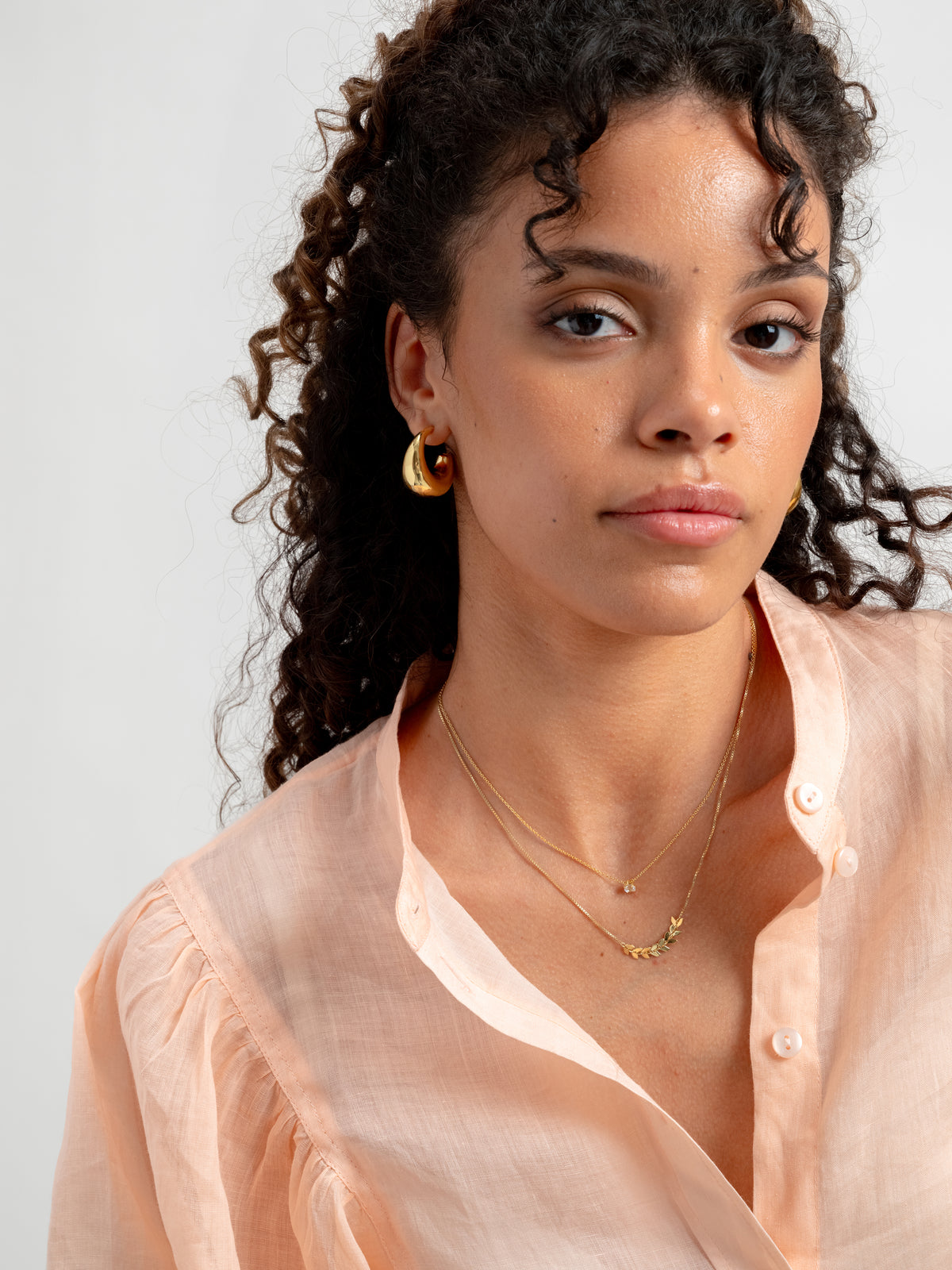 A model wears the Layers Simone Necklace in gold, featuring a delicate leaf pattern, layered with a simple thin gold chain and paired with chunky gold hoop earrings.