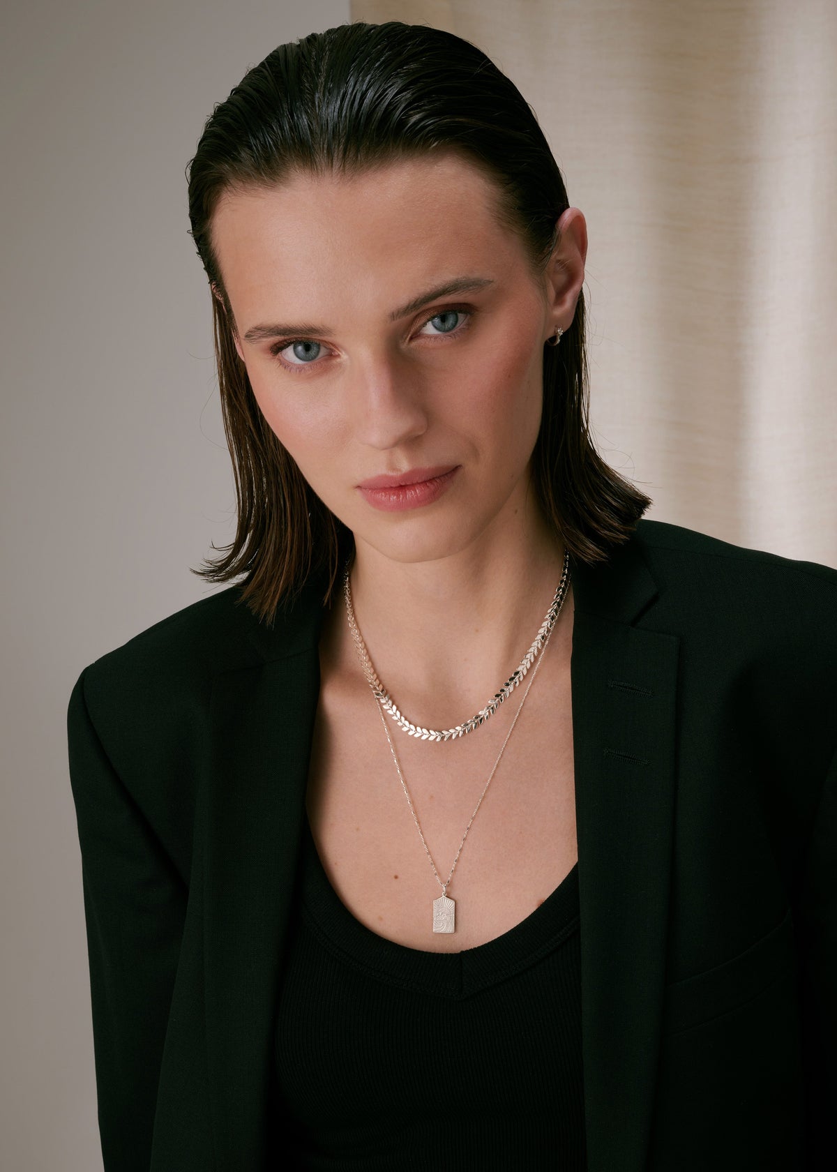 A model wearing two layered silver necklaces: a shorter one with a leaf pattern on top and a longer one with a rectangular pendant below. A small silver hoop earring is visible in her left ear.