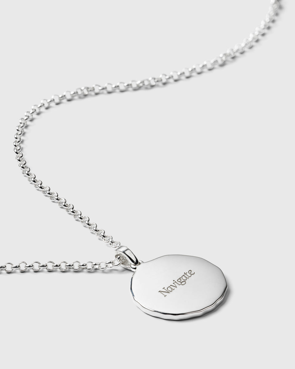 A close-up product shot of the Lucky Coin Navigate Necklace in silver, focusing on the back of the round pendant which is engraved with the word 'Navigate'.