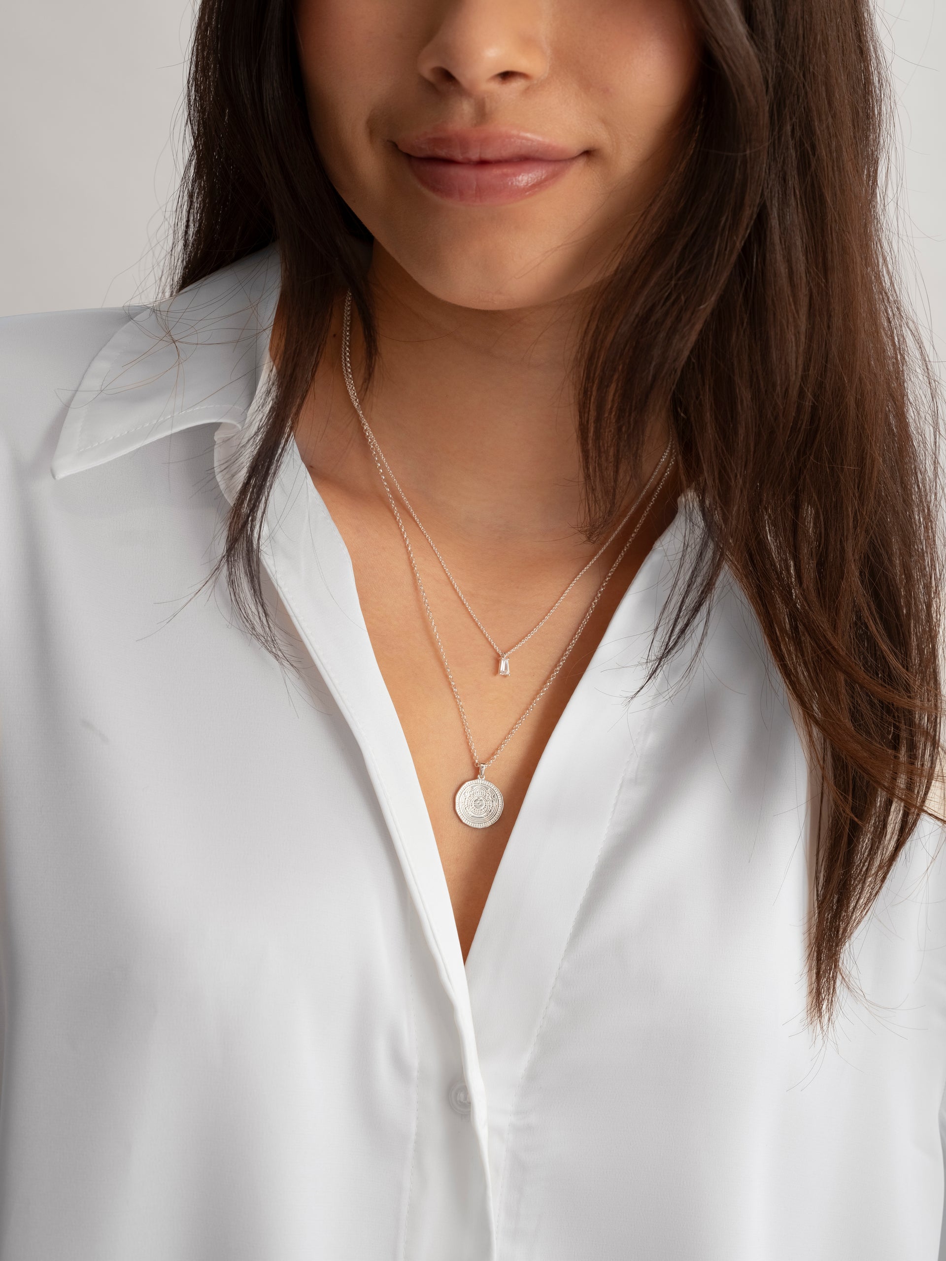 A model wears the silver Iggy Necklace, featuring a delicate chain and a single baguette-cut cubic zirconia, layered with a longer silver necklace that has an intricately engraved coin pendant.