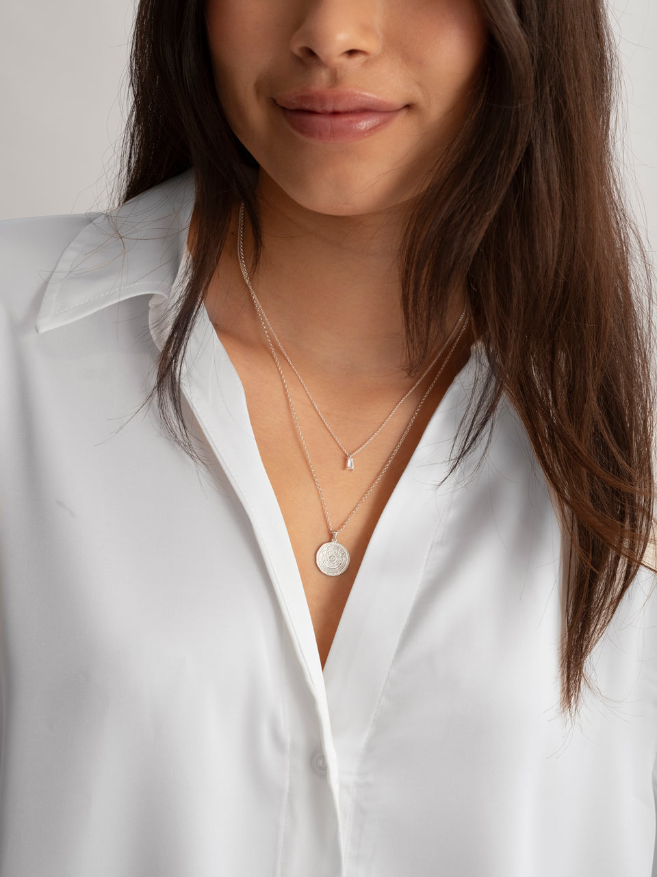 A model wears the silver Iggy Necklace, featuring a delicate chain and a single baguette-cut cubic zirconia, layered with a longer silver necklace that has an intricately engraved coin pendant.