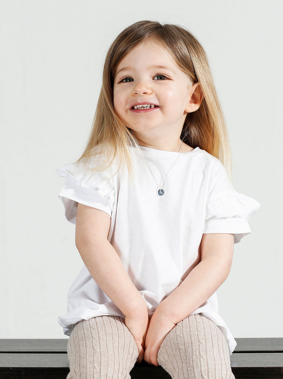 A young girl models the Beloved Baby Chain in silver, styled with a round blue pendant featuring the letter 'A'.