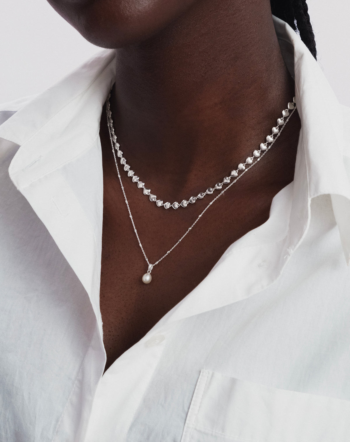 A close-up of a model wearing the Darling Necklace in silver, a chain made of interconnected hearts, layered with another fine silver chain featuring a small pearl pendant.