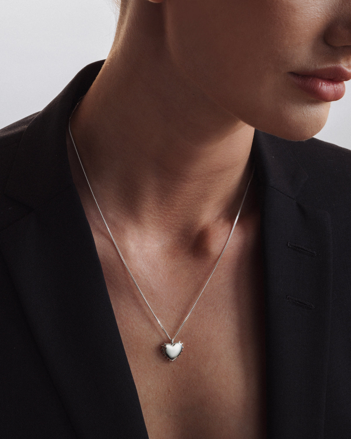 A close-up of a model's neck, wearing the True Love Spike Heart Necklace in silver, which features a chain and a heart pendant with small spikes, styled with a black blazer.