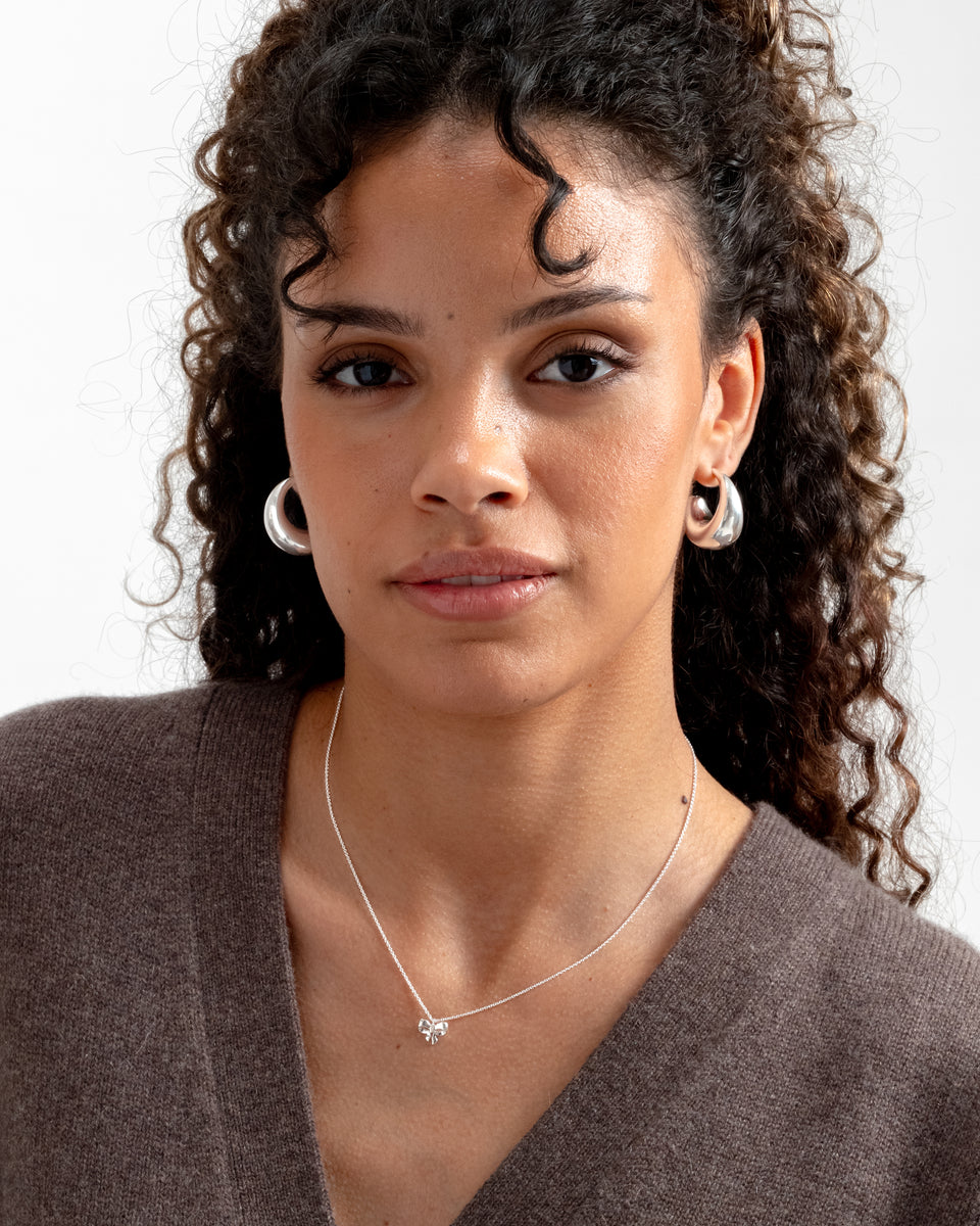 A model wears the Rosie Mini Necklace in silver, featuring a delicate bow pendant, styled with a pair of chunky silver hoop earrings.