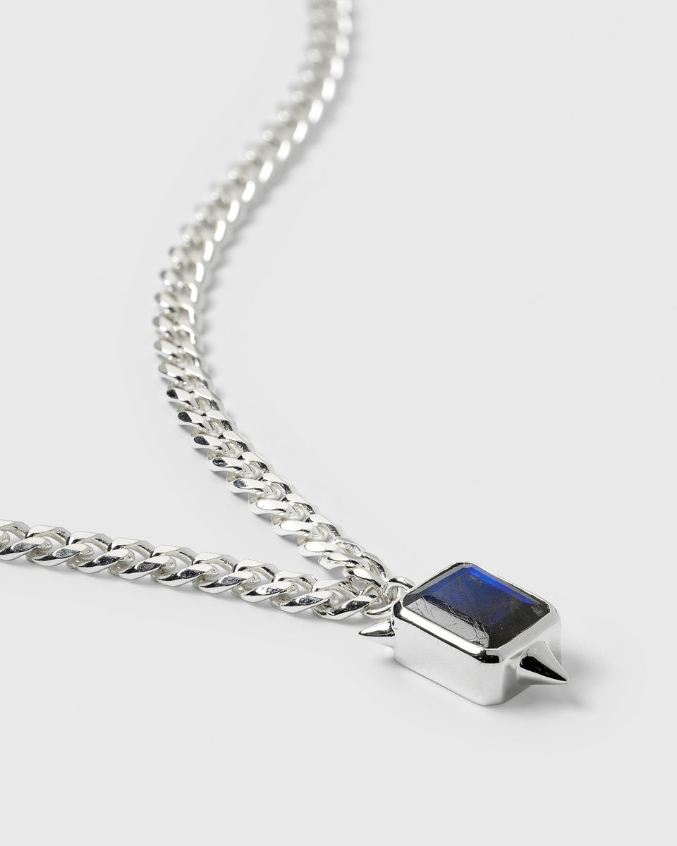 A product shot of the Drama Necklace in silver, showcasing its heavy chain and the rectangular pendant set with a labradorite stone and adorned with spikes.