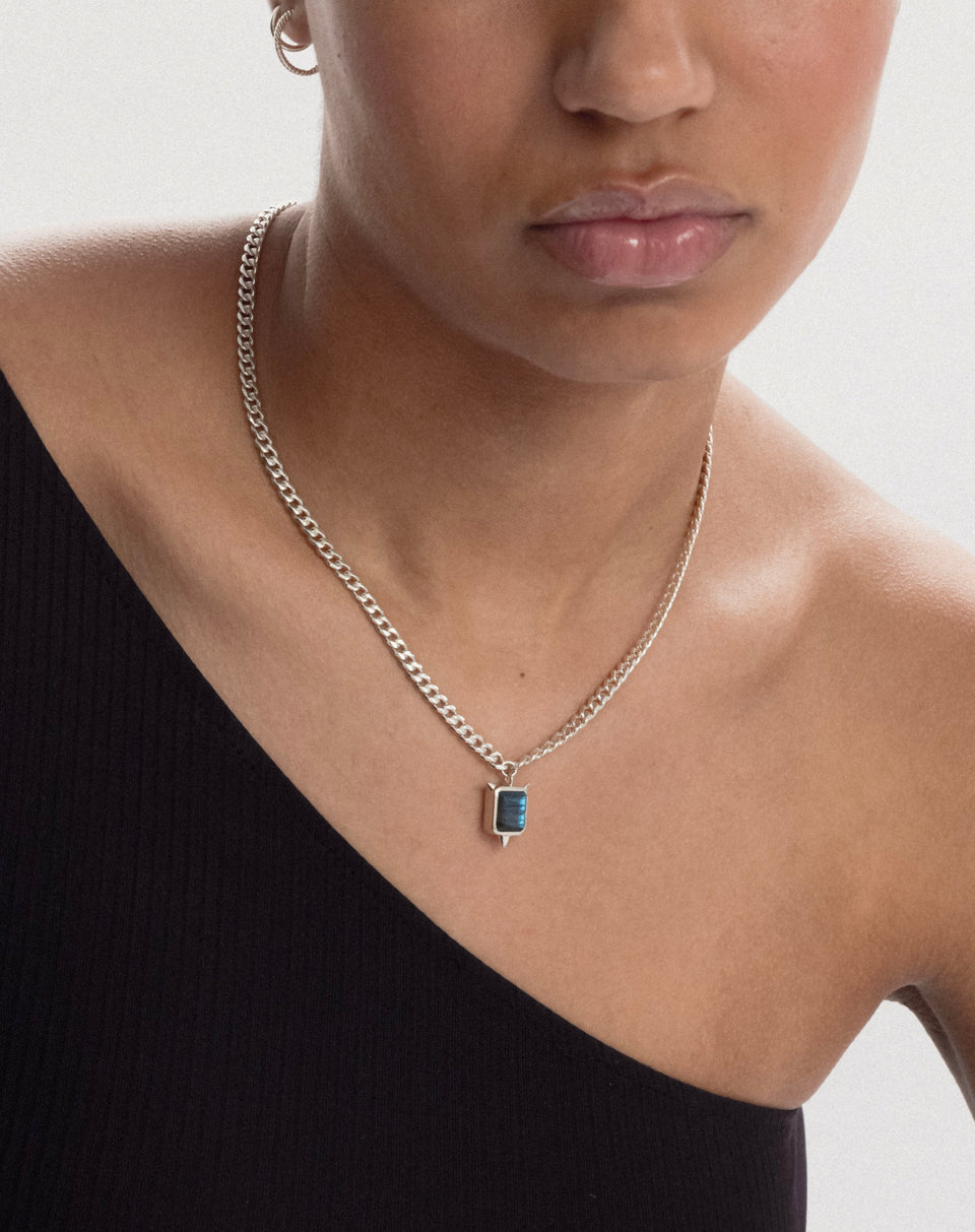A close-up of a model wearing the Drama Necklace in silver, which features a curb chain and a rectangular labradorite pendant with spikes on its corners. She is also wearing a small silver hoop earring.
