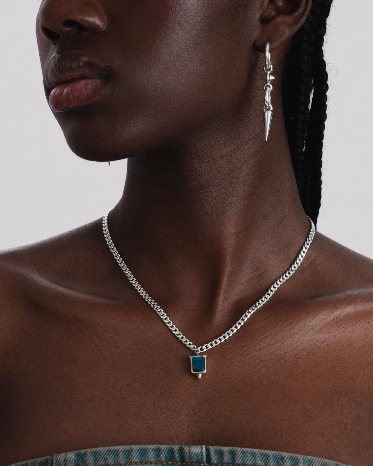 A close-up of a model wearing the Drama Necklace in silver, featuring a heavy chain and a rectangular labradorite pendant with spikes, paired with a matching silver spike earring.