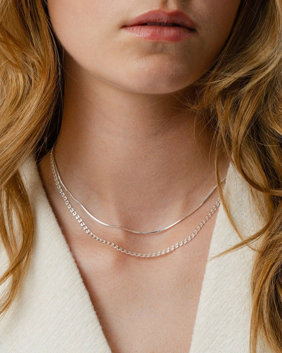 A close-up shot of a model wearing the Sheer Mirror Necklace in silver, a smooth snake chain, layered over a thicker silver curb chain necklace.