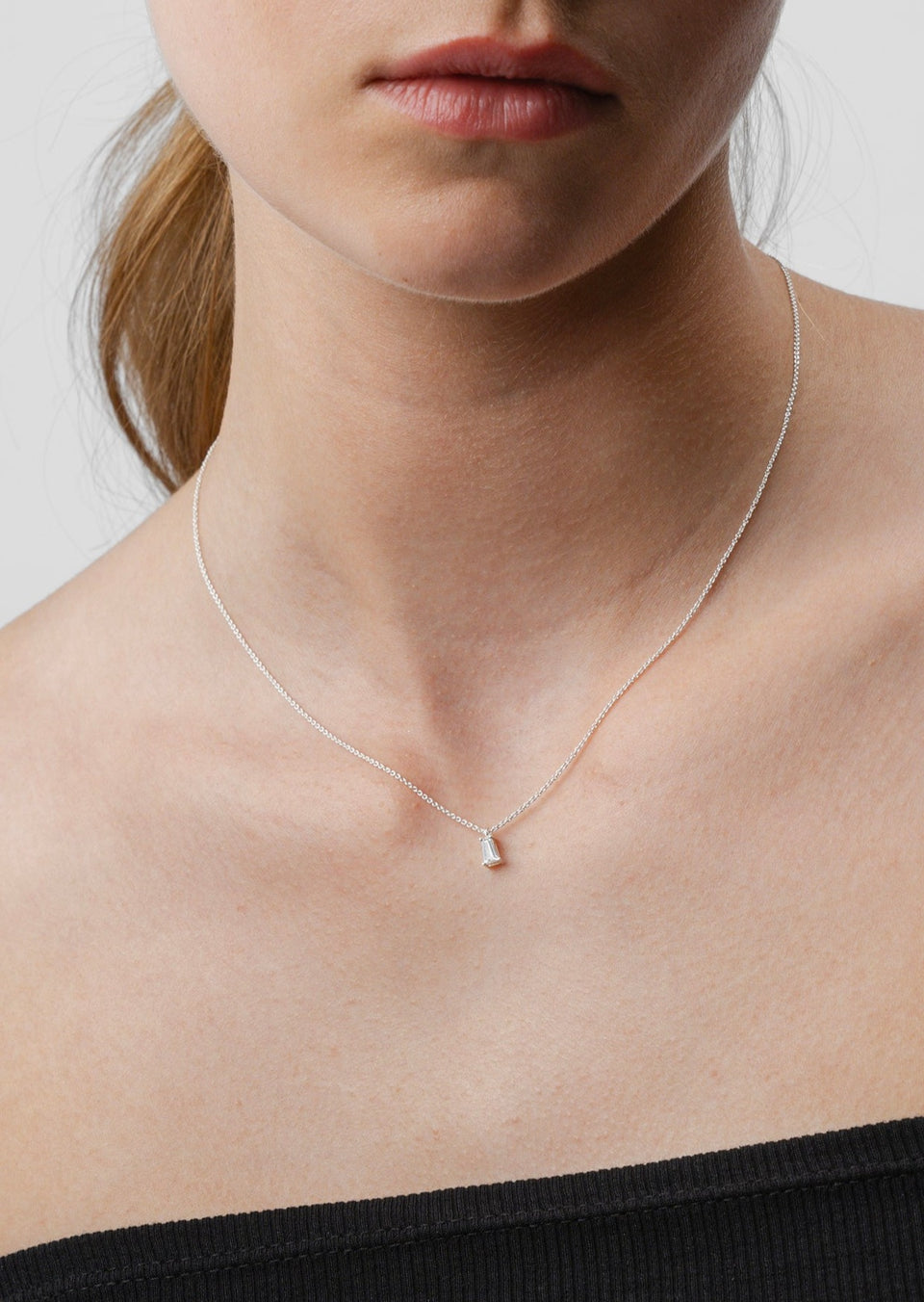 A close-up shot of a model wearing the Iggy Necklace in silver, featuring a delicate chain and a single tapered baguette-cut cubic zirconia pendant.