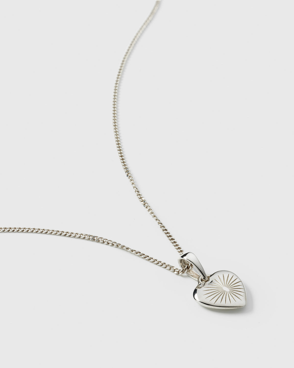 A product shot of the Sunburst Heart Necklace in silver, featuring a fine chain and a heart pendant with an engraved sunburst pattern.