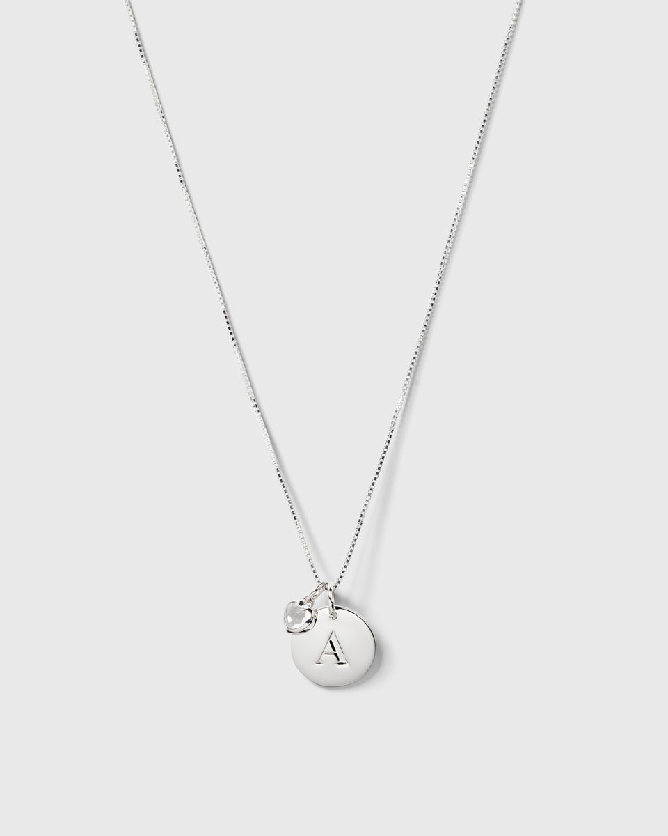 A product shot of a silver necklace featuring two pendants: the small, crystal Beloved Heart Pendant and a circular silver disc engraved with the letter 'A'.