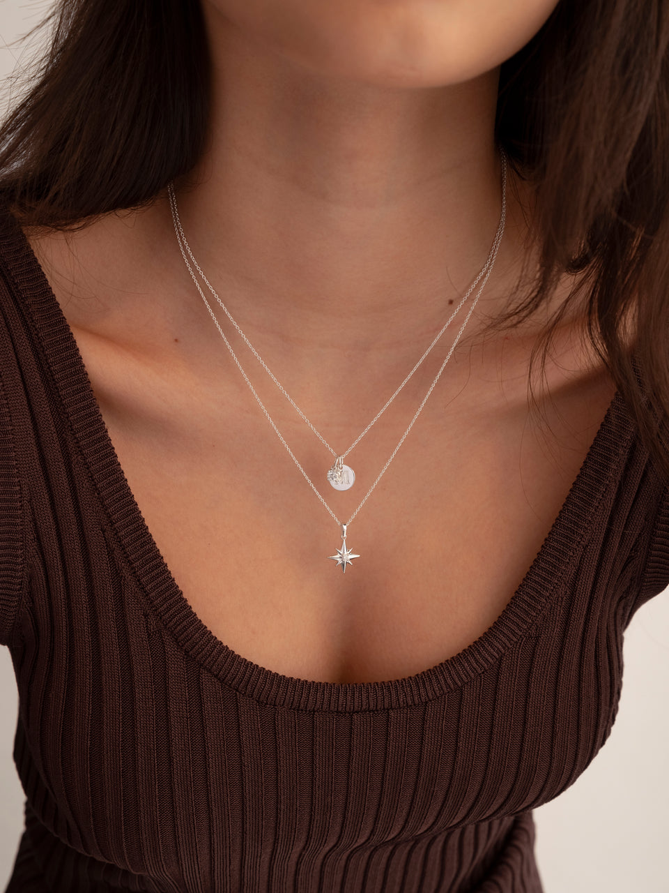 A model wearing the North Star Big Necklace in silver, featuring a star-shaped pendant, layered with a second silver necklace with an initial 'M' pendant.