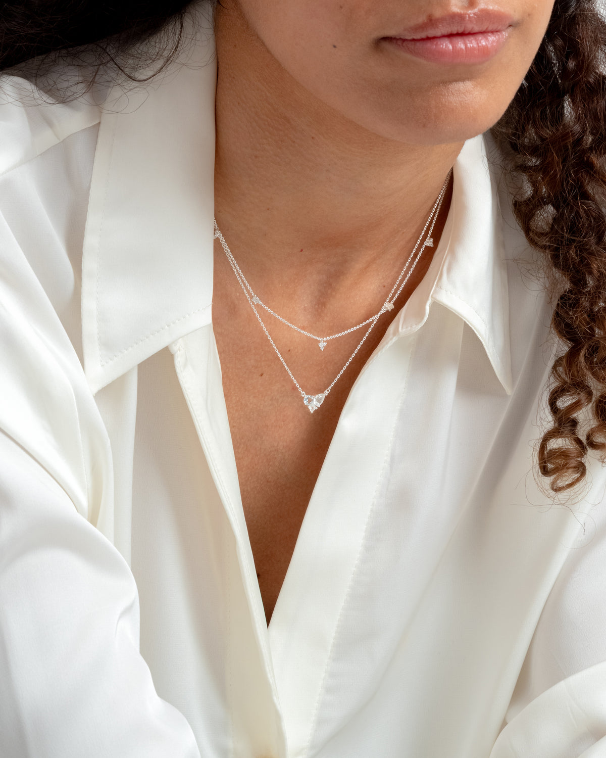 A close-up of a model wearing the Celestia Necklace in silver, layered with a longer silver necklace with a heart-shaped stone, against her neck and a white shirt.