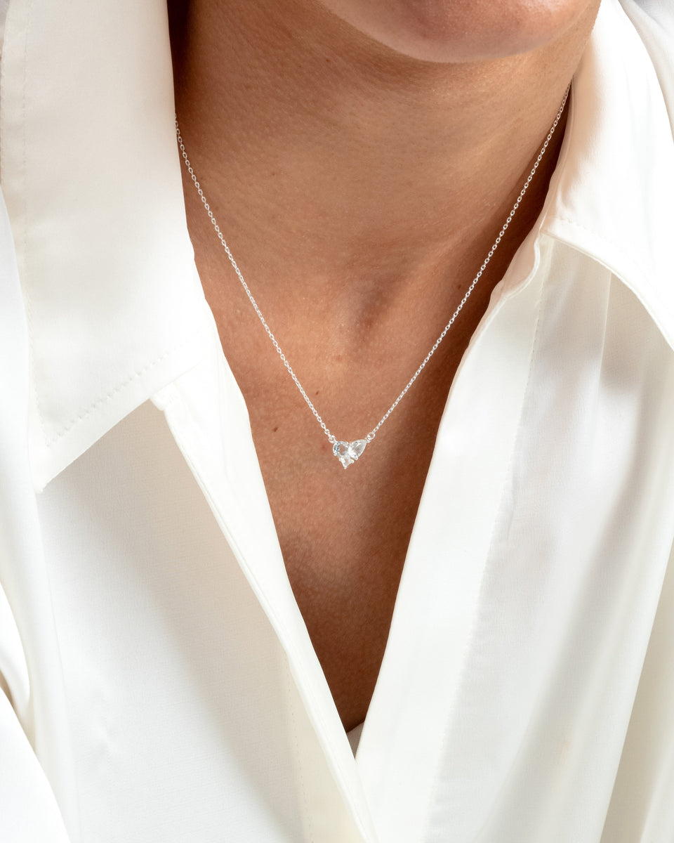 A close-up of a model wearing the Pam Necklace in silver, styled with a white shirt. The necklace features a delicate chain and a pendant with three sparkling, clustered white topaz stones.