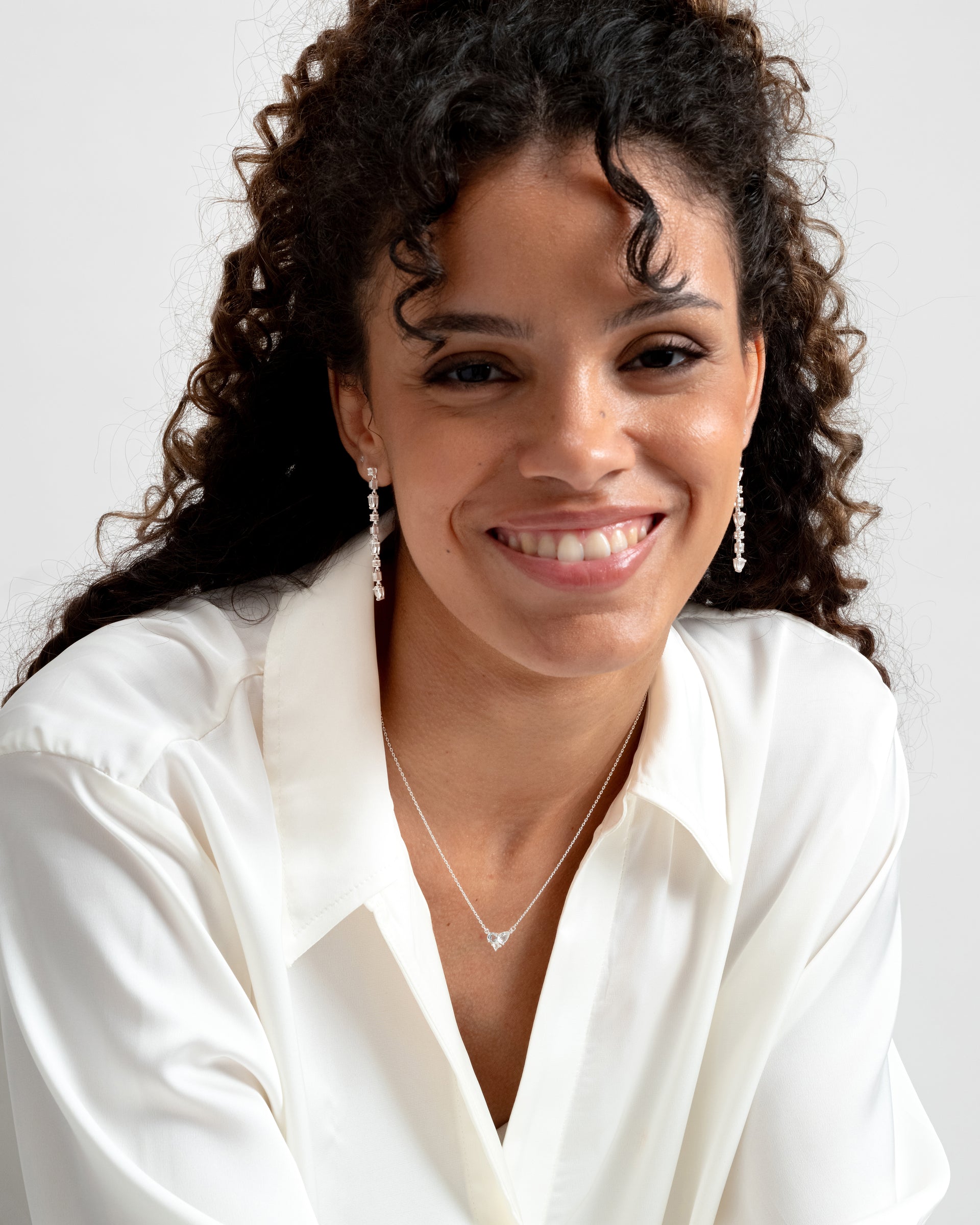 A smiling model wears the Pam Necklace in silver, featuring a delicate chain and a sparkling white topaz pendant, paired with matching drop earrings.