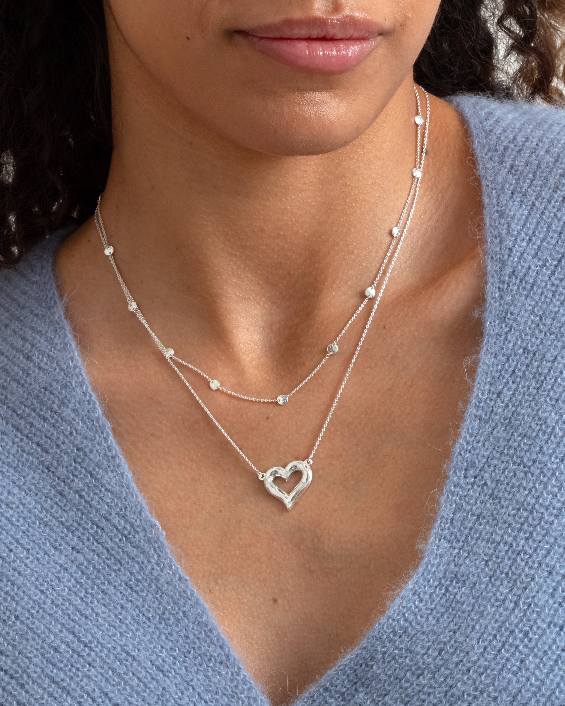 A close-up of a model wearing the silver Solea Necklace with its hammered circles details, layered with a second silver necklace featuring a hammered open-heart pendant, styled with a blue knit sweater.