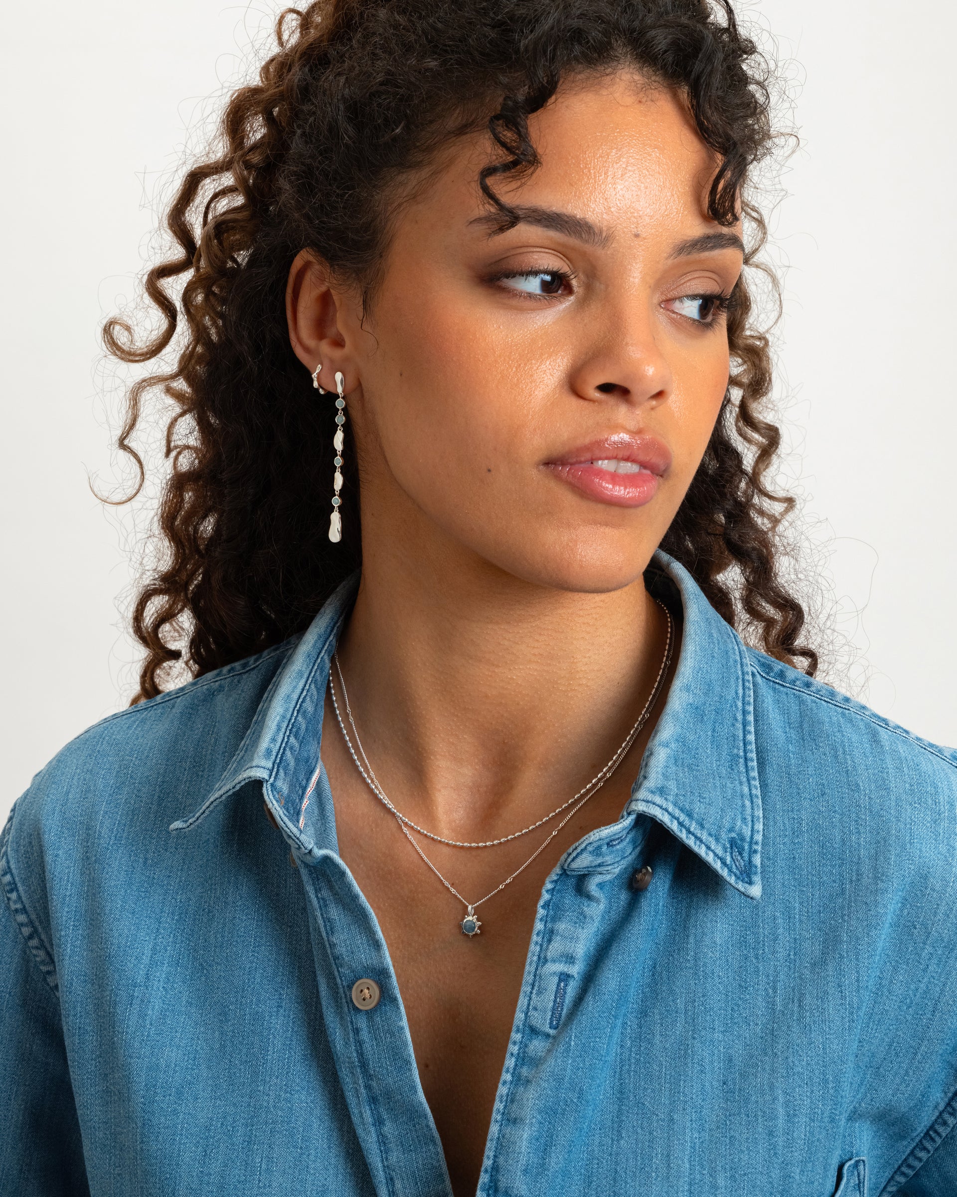 A model with curly hair wearing the Lunetta Necklace in silver, featuring a ball chain with oval beads. It is layered with a finer silver necklace with a small light blue pendant, and paired with dangling silver pearl earrings.