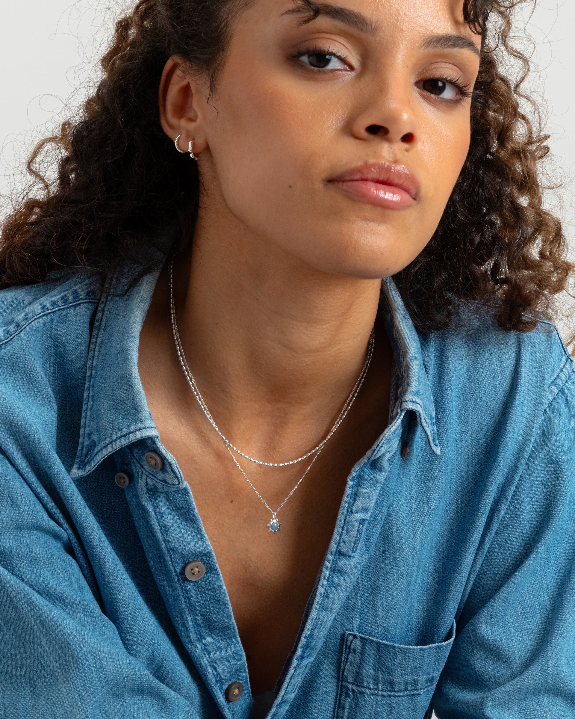A model wears the silver Lunetta ball chain necklace layered with a delicate silver chain necklace with a light blue stone pendant, paired with small silver hoop earrings.