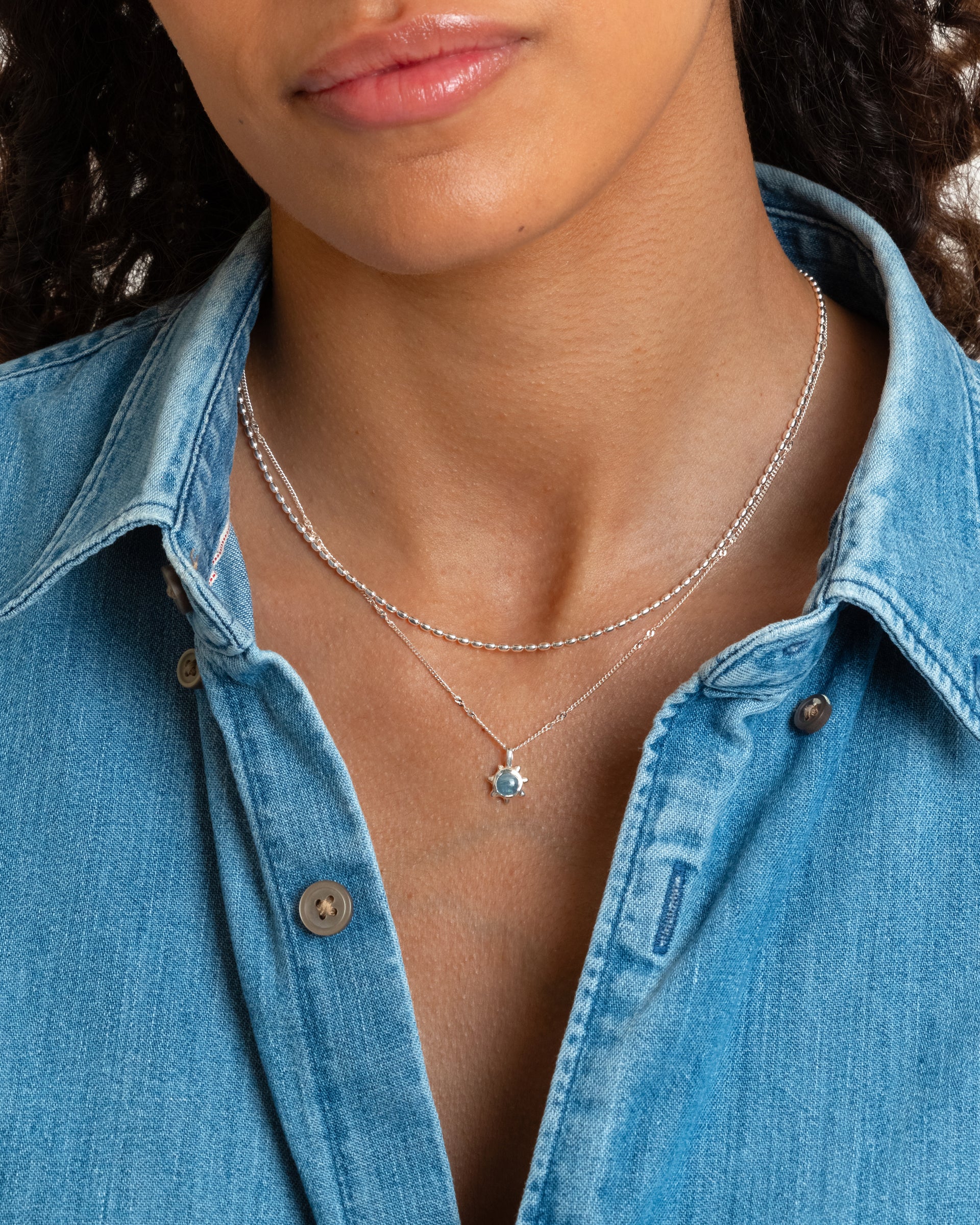 A model wears the Lunetta Necklace in silver, a ball chain with oval beads, layered with a delicate silver chain necklace featuring a sun-shaped pendant with a light blue stone against a denim shirt.