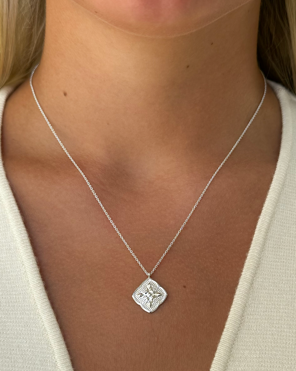 A close-up of a model wearing the Luna Necklace in silver, showcasing the fine chain and the intricately detailed, quatrefoil-shaped pendant.