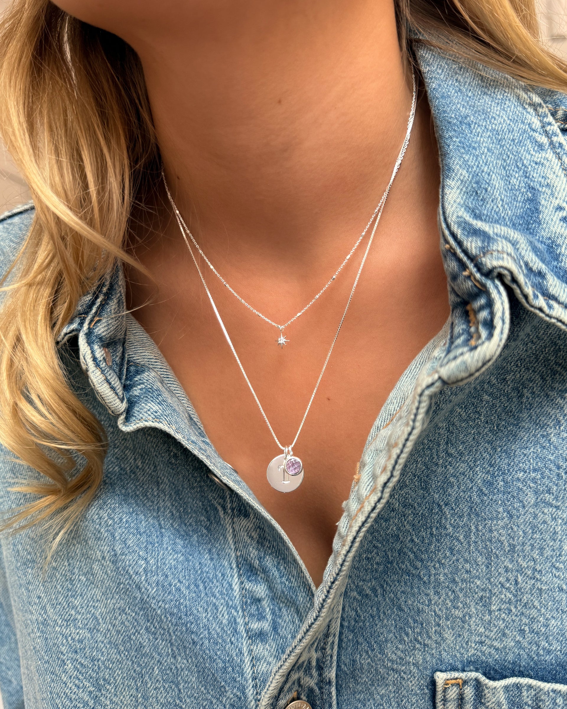 Two layered silver necklaces: a longer chain featuring a February birthstone amethyst pendant and a round letter charm, paired with a shorter necklace with a delicate star pendant.