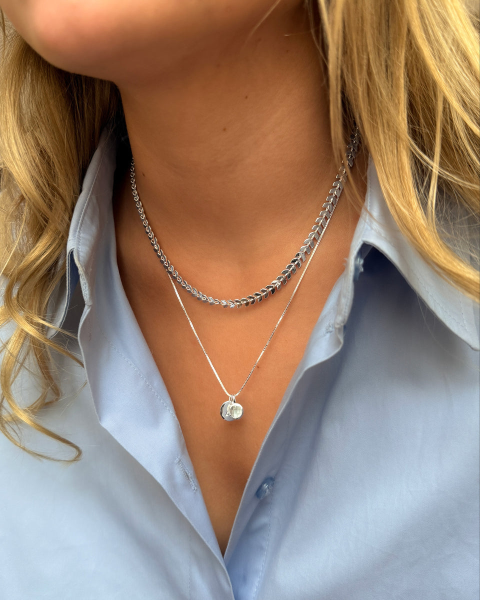 A silver chain featuring a March birthstone pendant with a light blue aquamarine stone, layered with a second silver chain adorned with a delicate leaf design.