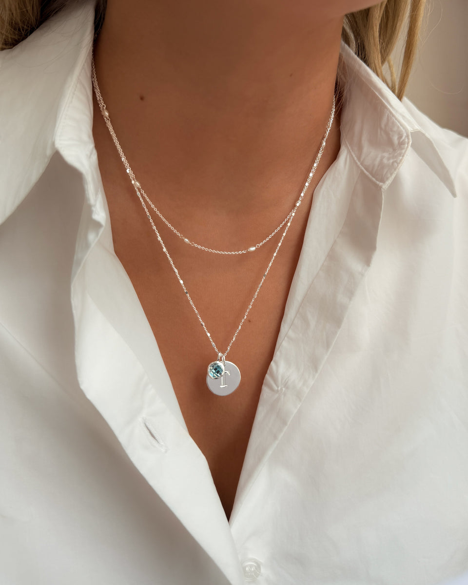 Two layered silver necklaces with a longer chain featuring a September birthstone blue topaz pendant and a round letter charm.
