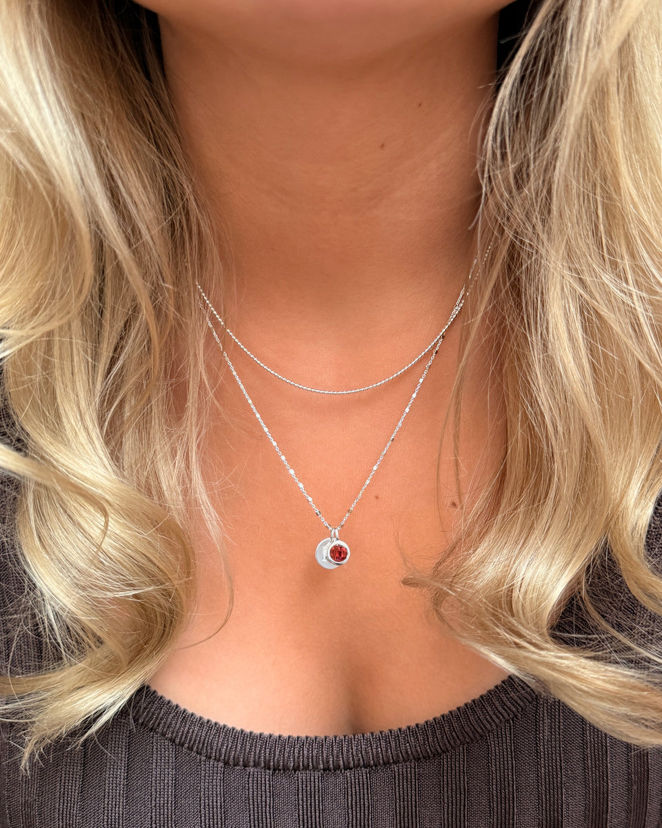 A silver pendant necklace featuring a round January birthstone garnet, layered with a simple silver chain