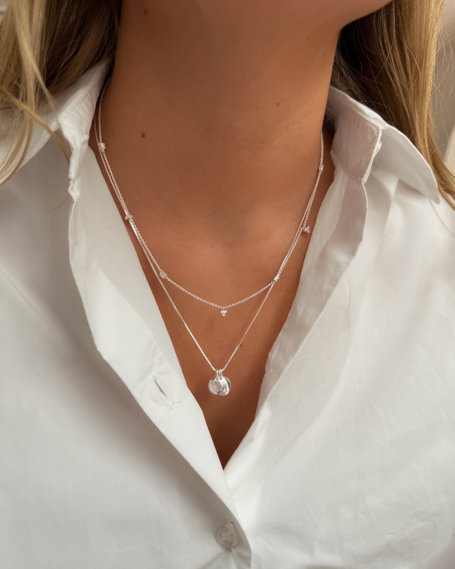 Two layered silver necklaces: the longer necklace with a June birthstone moonstone pendant and a small round silver letter pendant, and the shorter necklace with a delicate chain and a small crystal pendant.