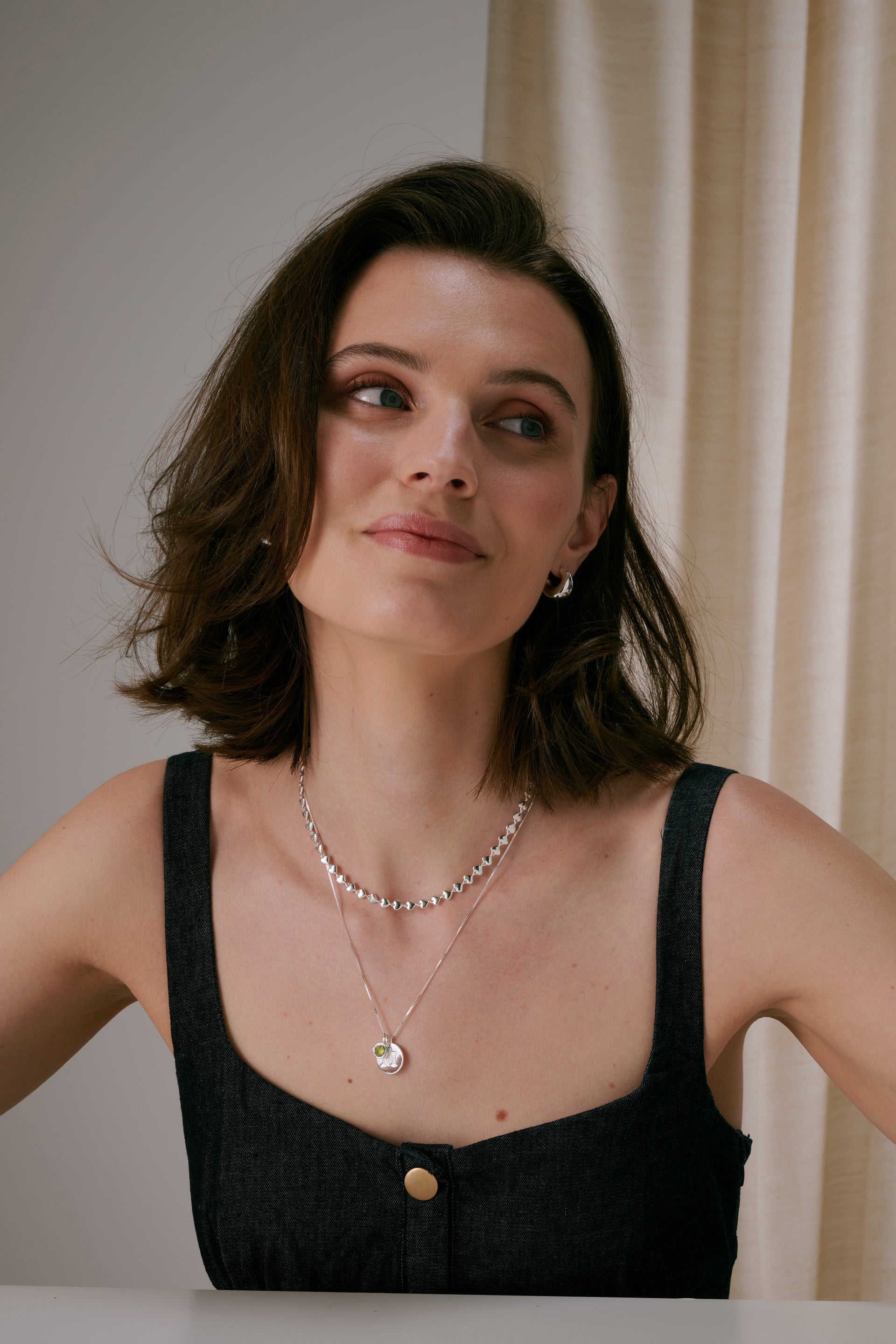 A pair of silver hoop earrings paired with two layered silver necklaces. The shorter necklace features a heart-decorated pattern, while the longer chain holds a large round letter pendant and a light green August birthstone charm.