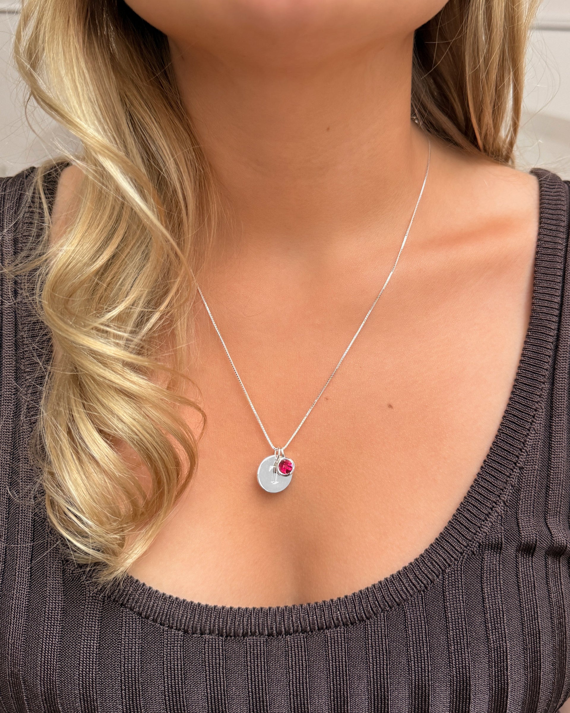 A silver necklace with a ruby pendant, July’s birthstone, layered with a delicate silver chain featuring a small letter pendant.