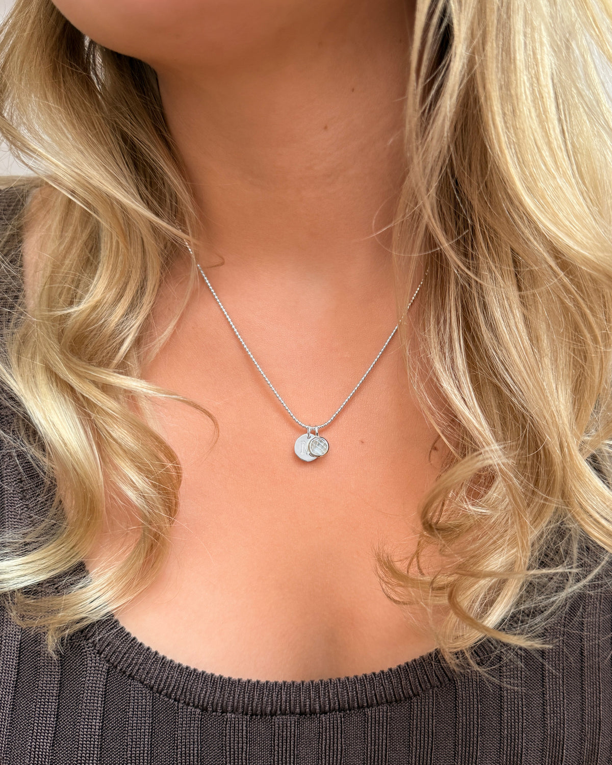 A silver pendant featuring the April birthstone, white topaz, worn on a silver chain with a round letter charm.