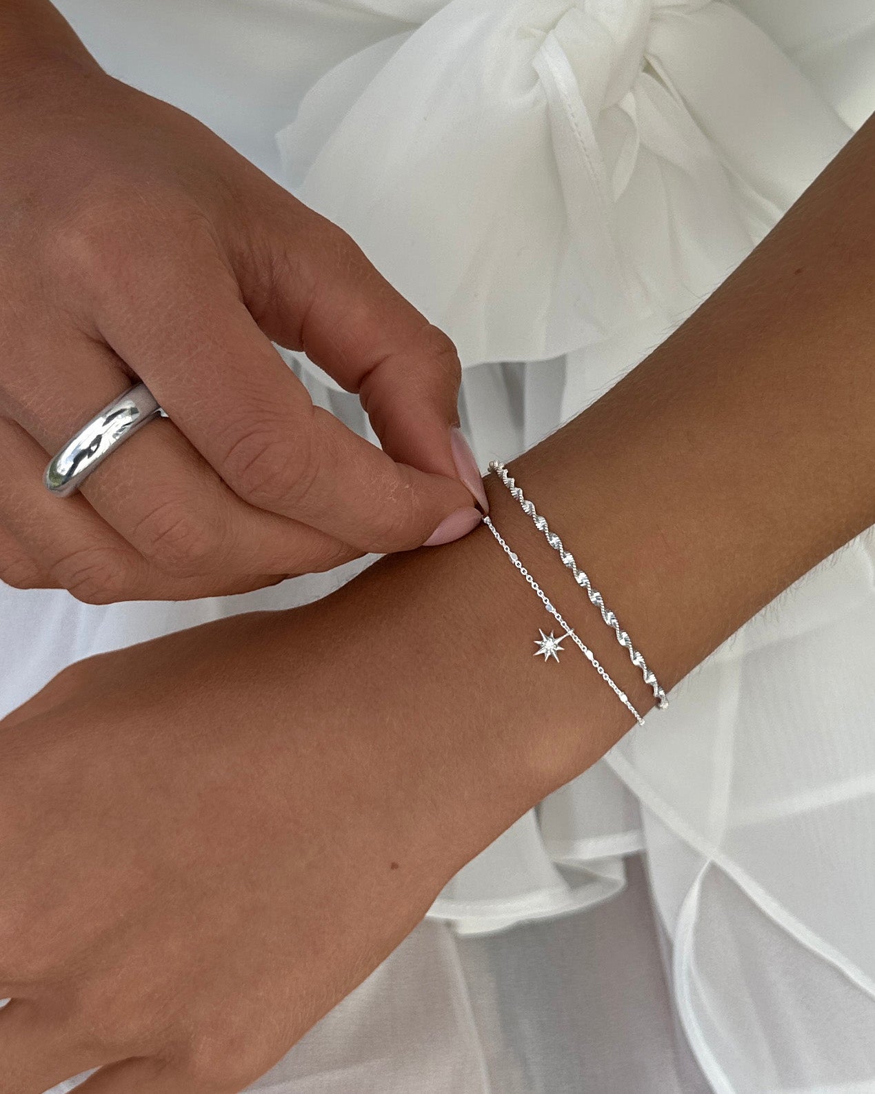 A close-up of a model adjusting the silver North Star Bracelet, featuring a delicate star charm. It's layered on her wrist with a Herringbone Twisted Bracelet in silver. She also wears a bold silver ring.