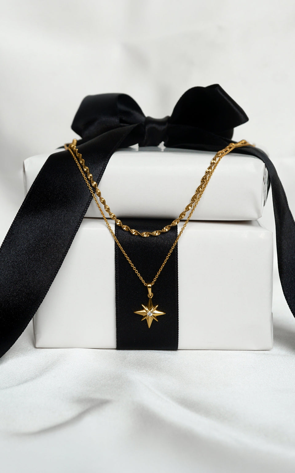 A product shot of the North Star & Herringbone Necklace Set in gold, presented on a white gift box with a black ribbon, showcasing the layered look of the twisted herringbone chain and the fine chain with a North Star pendant.
