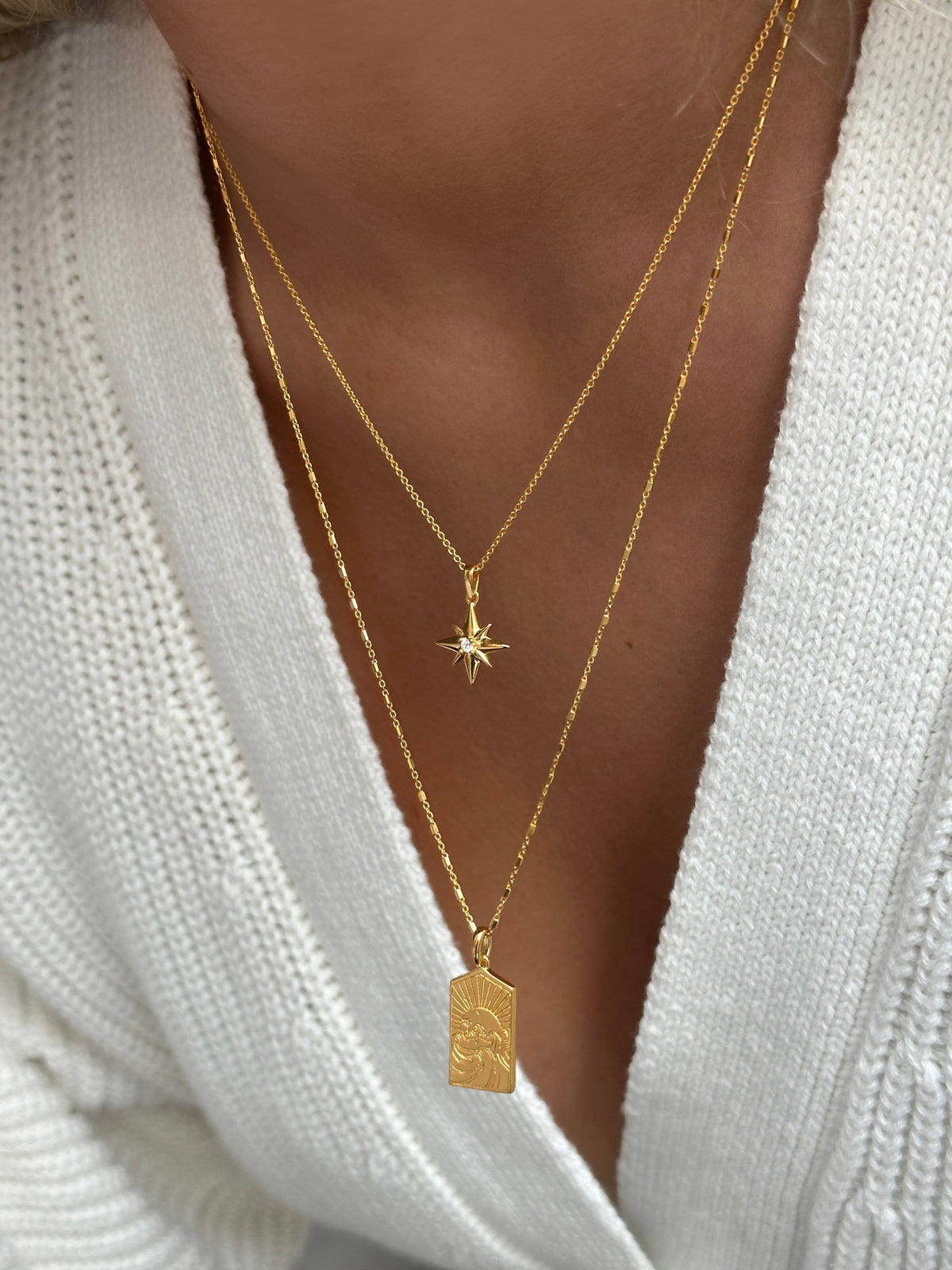 A close-up of a model wearing a white knitted sweater, styled with two layered gold necklaces. The longer necklace is the Lucky Coin Keep Dreaming Necklace with a rectangular pendant, layered with the shorter North Star Necklace featuring a star-shaped pendant.