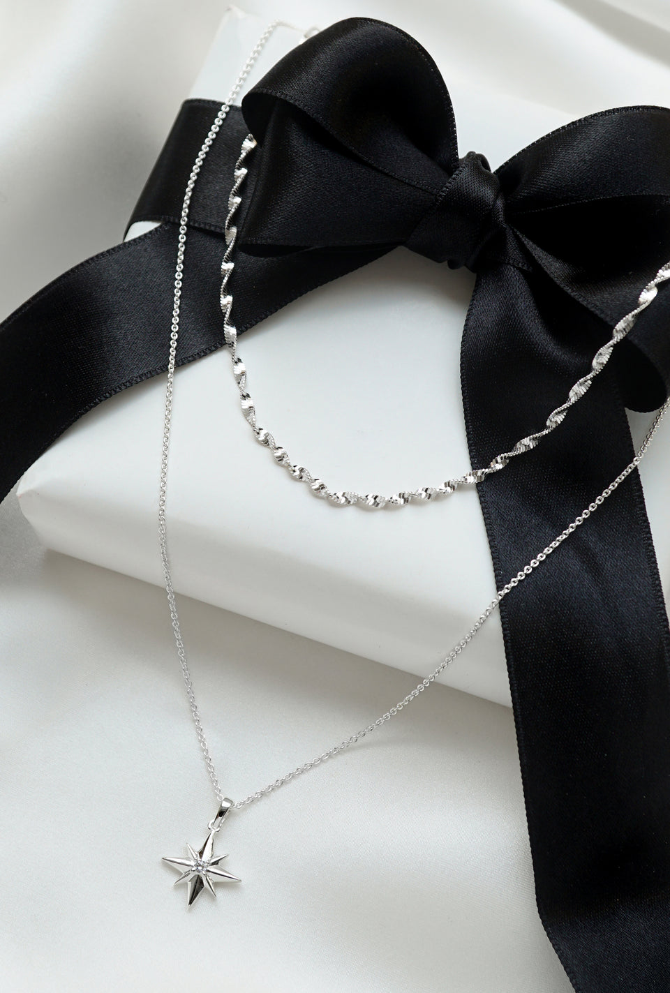 A product shot of the North Star & Herringbone Necklace Set in silver, displayed on a white gift box with a black ribbon, showing both the twisted herringbone chain and the North Star pendant necklace.