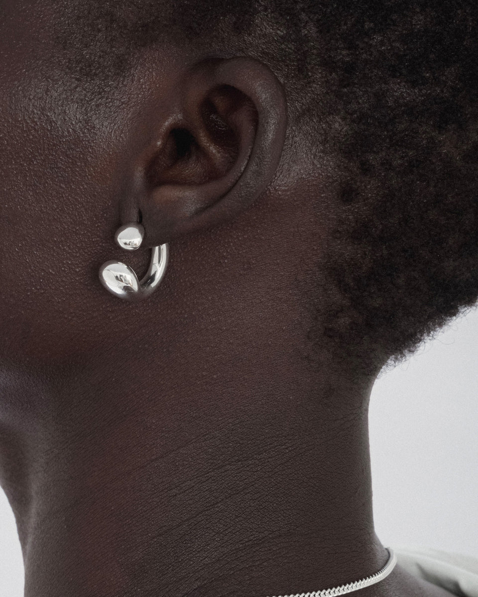A close-up of a model wearing the Bolded Drops Earrings in silver, showcasing their unique design with two drop shapes, and a thin silver chain necklace.