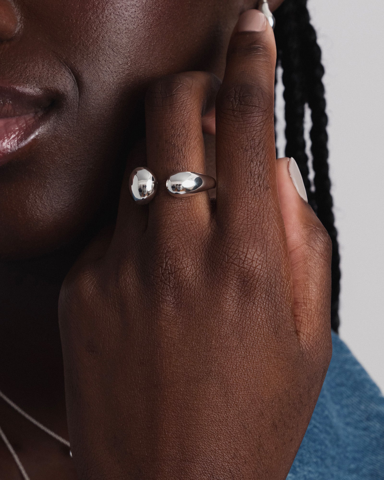 A close-up of a model wearing the Bolded Drops Ring Silver, highlighting its bold, open design with two polished drop shapes on her finger. A fine silver necklace is also visible in the background.