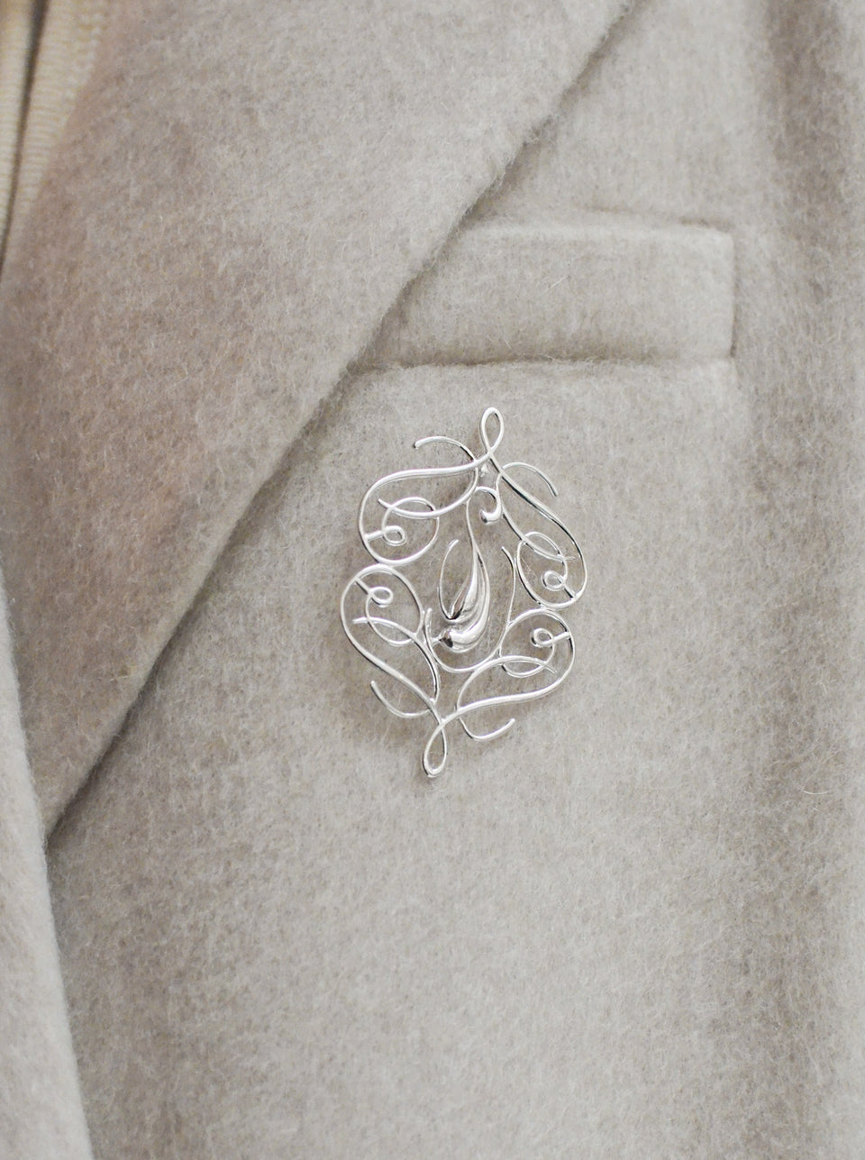 A shot of the Ophelia Brooch in silver, featuring an elegant swirling pattern, pinned to the lapel of a light-colored coat.