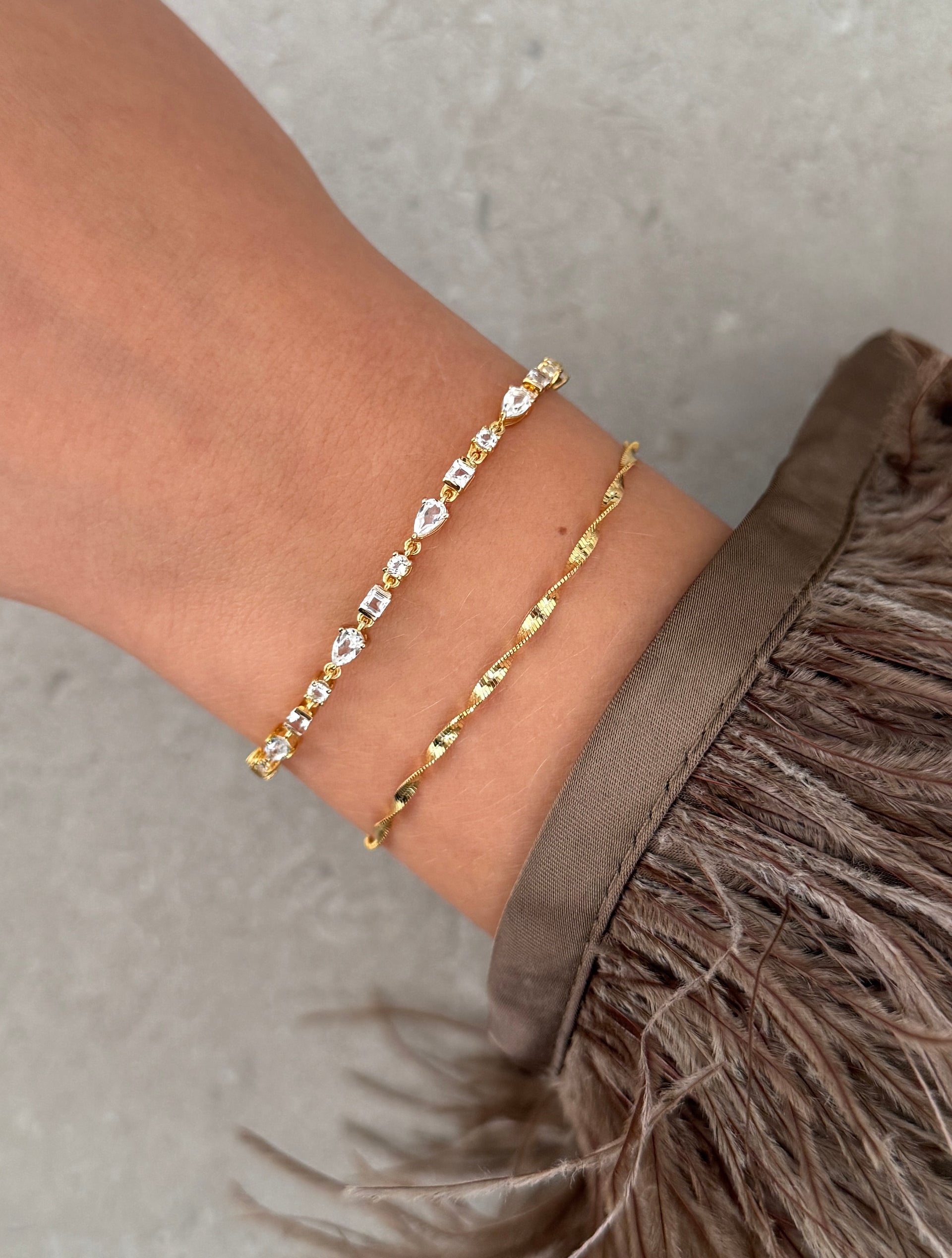 A close-up shot of a model's wrist wearing the Herringbone Twisted Bracelet in gold, shown stacked with a delicate gold and clear stone bracelet.